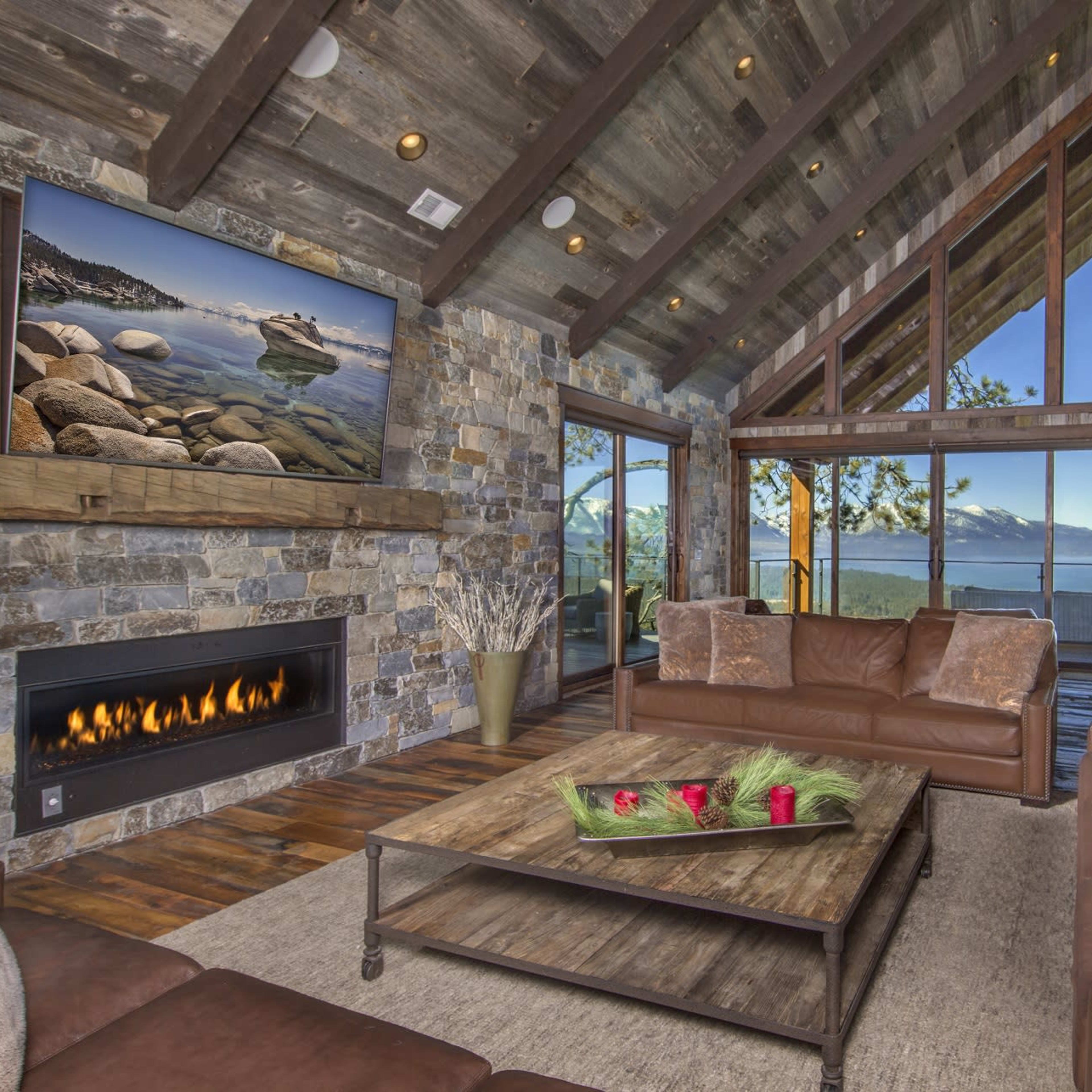 The living room features a stone fireplace, large windows with a view of the mountains, and leather furniture arranged around a coffee table.