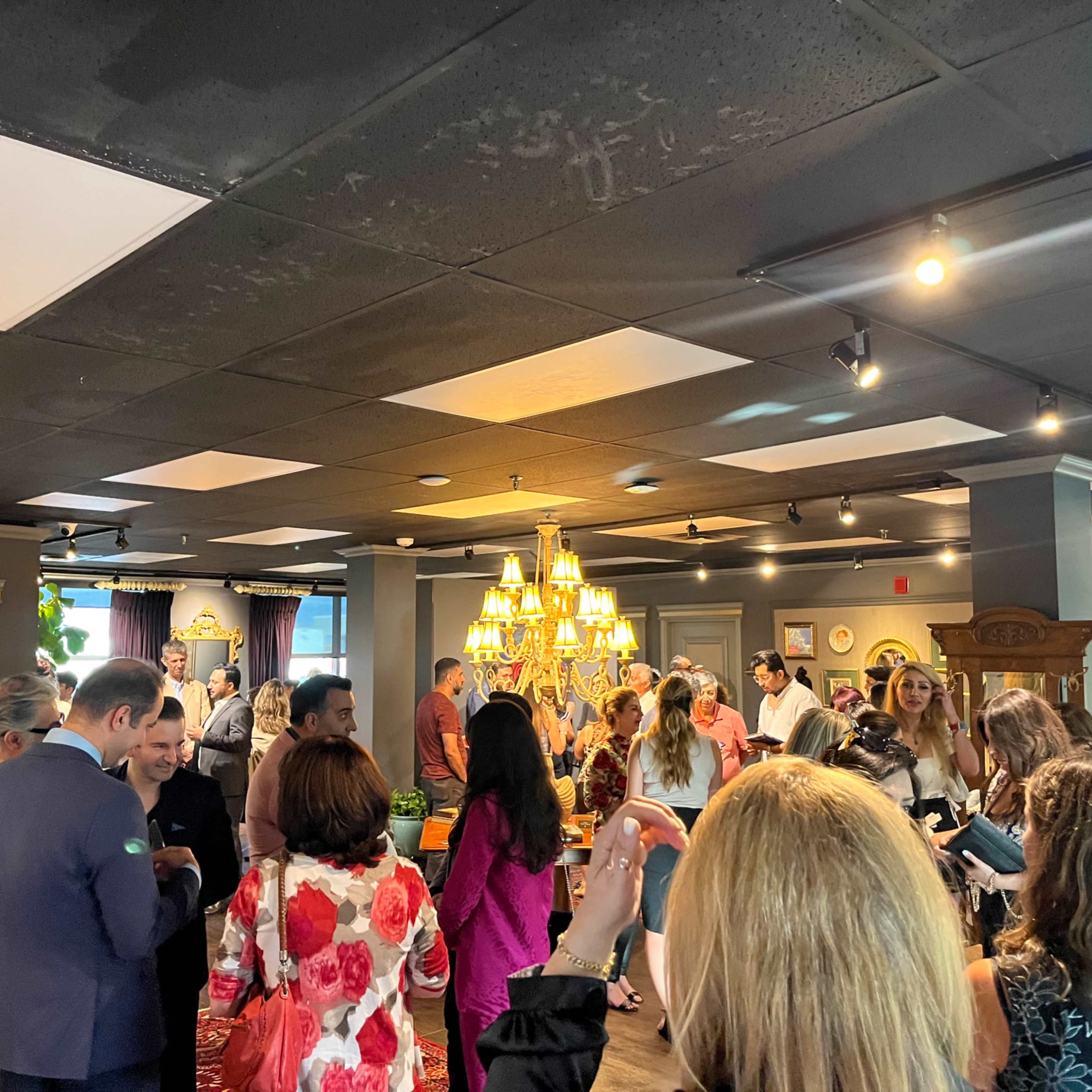 A gathering of people is seen in a well-lit indoor setting, featuring a chandelier and various decorative elements.