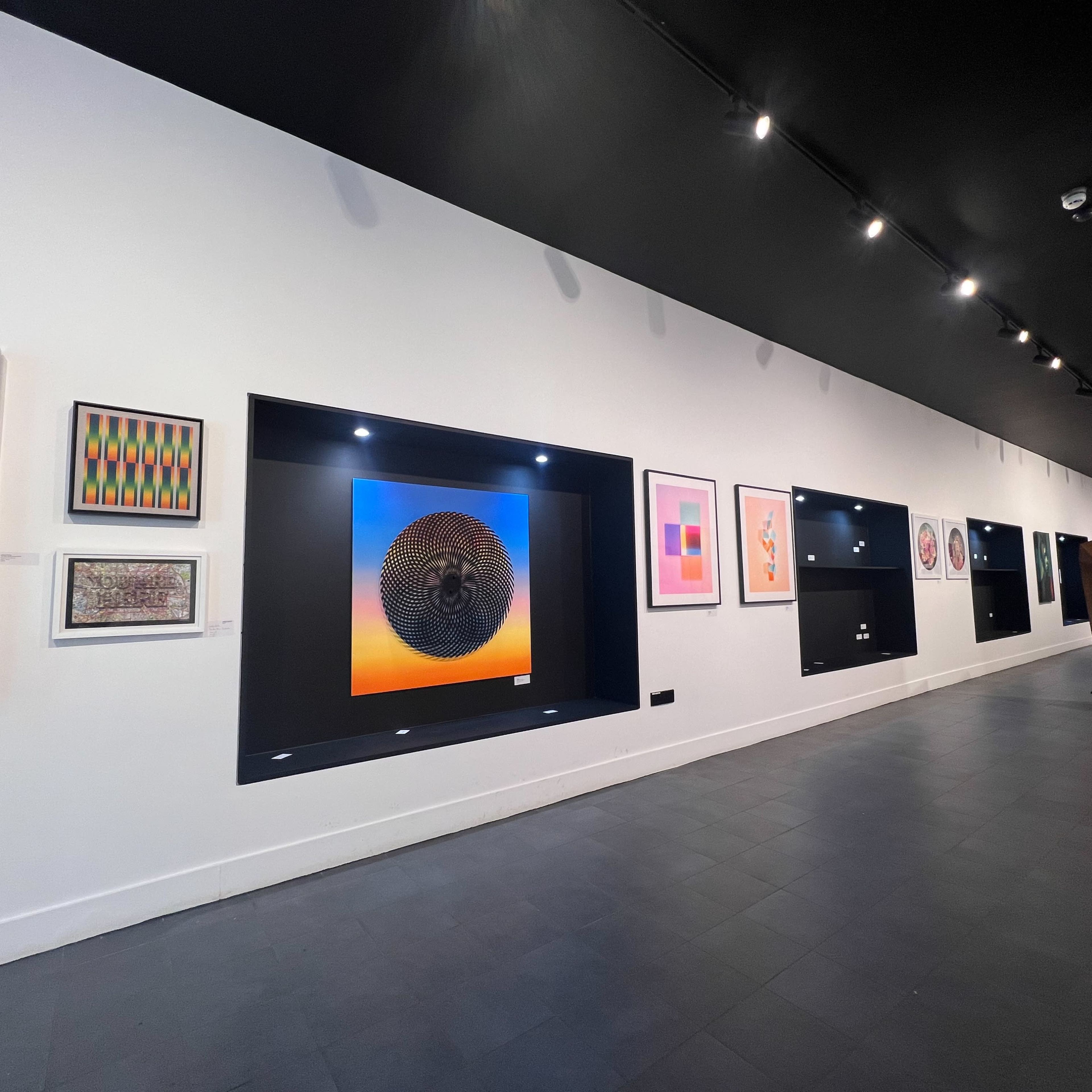 An interior of an art gallery with various framed artworks displayed on a white wall and a large sculptural piece on the right.