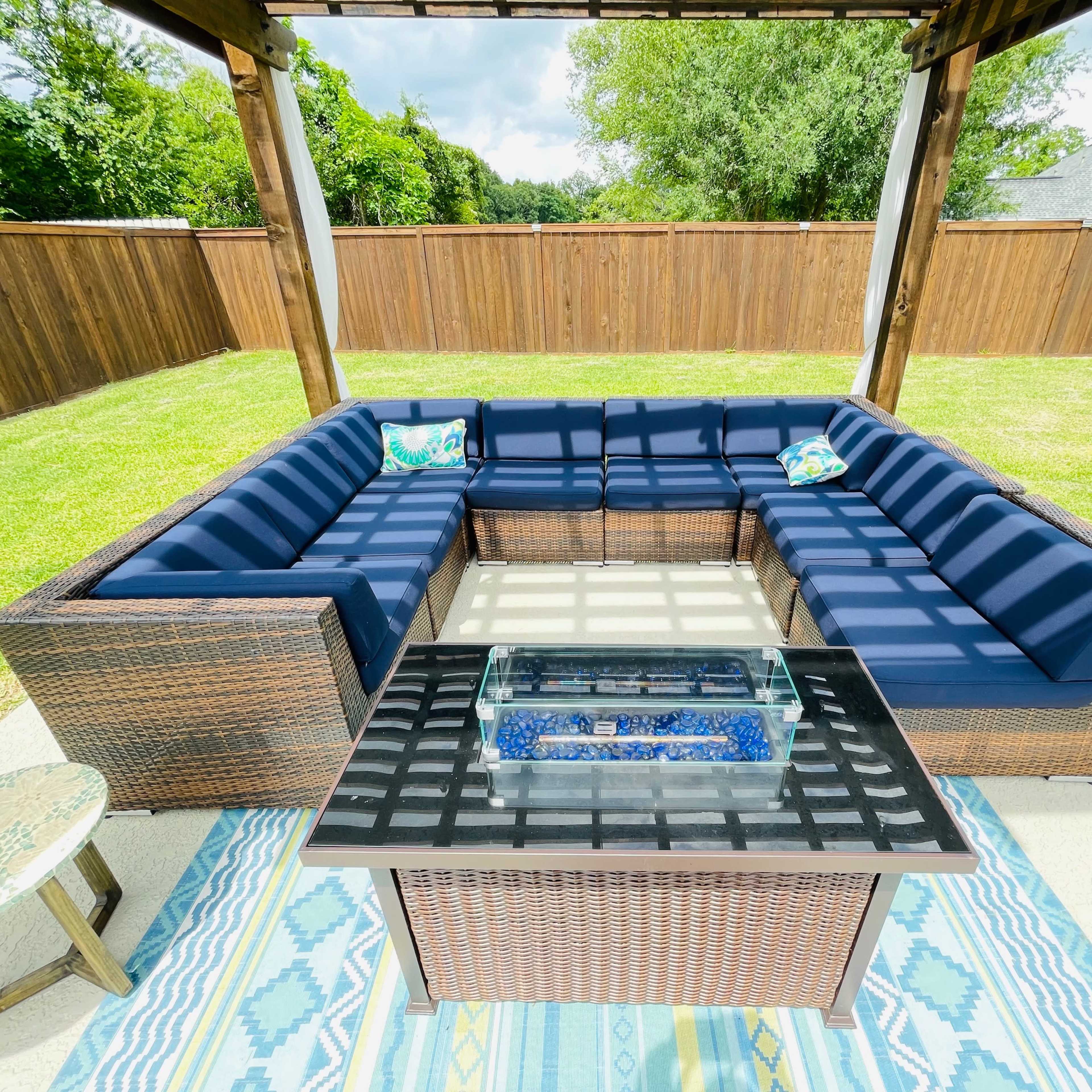 A spacious outdoor seating area features a large sectional couch with navy cushions and a glass-top fire pit table, all set on a patio surrounded by a grassy yard and wooden privacy fence.