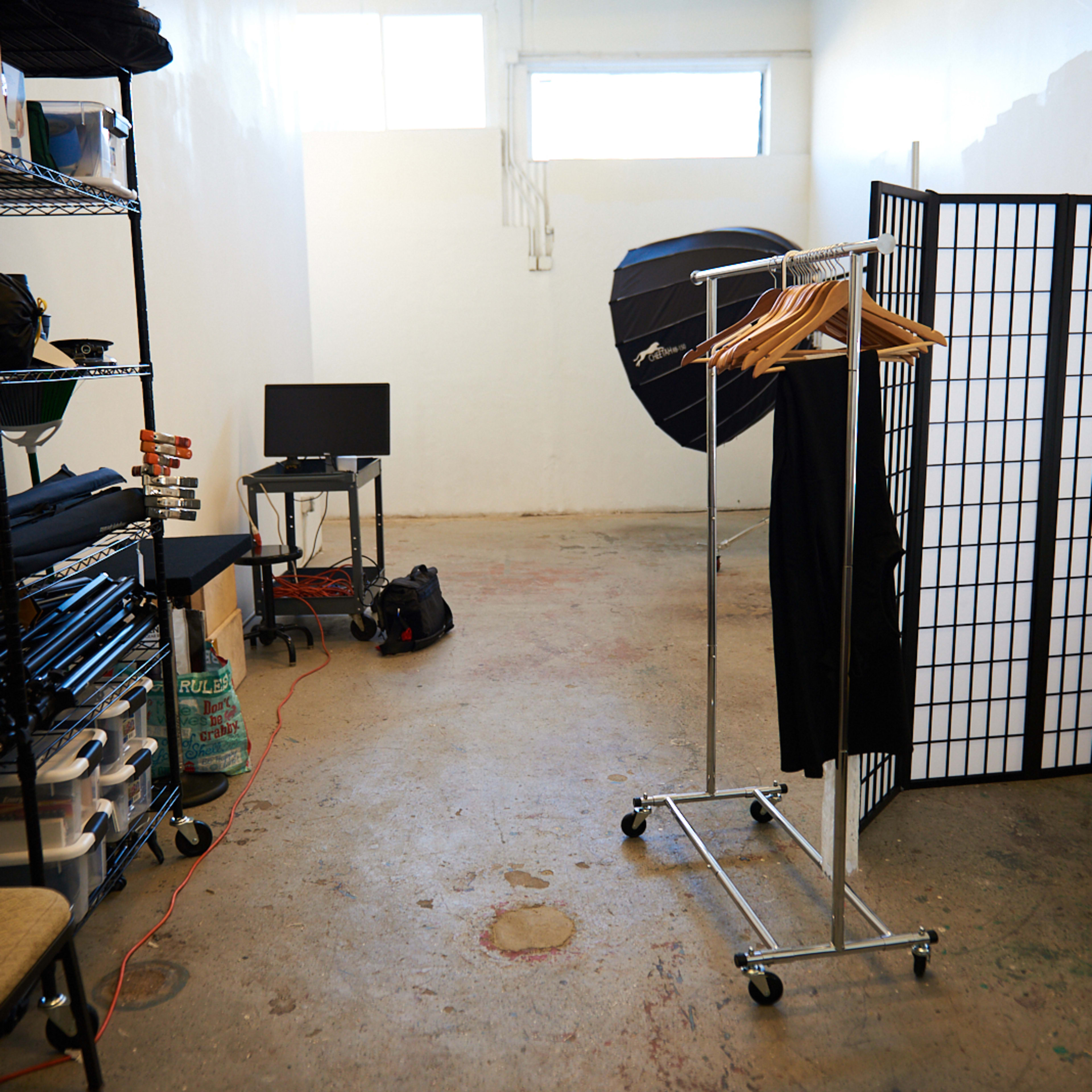 An empty studio space features a clothing rack, a monitor on a table, folding chairs, and a room divider.