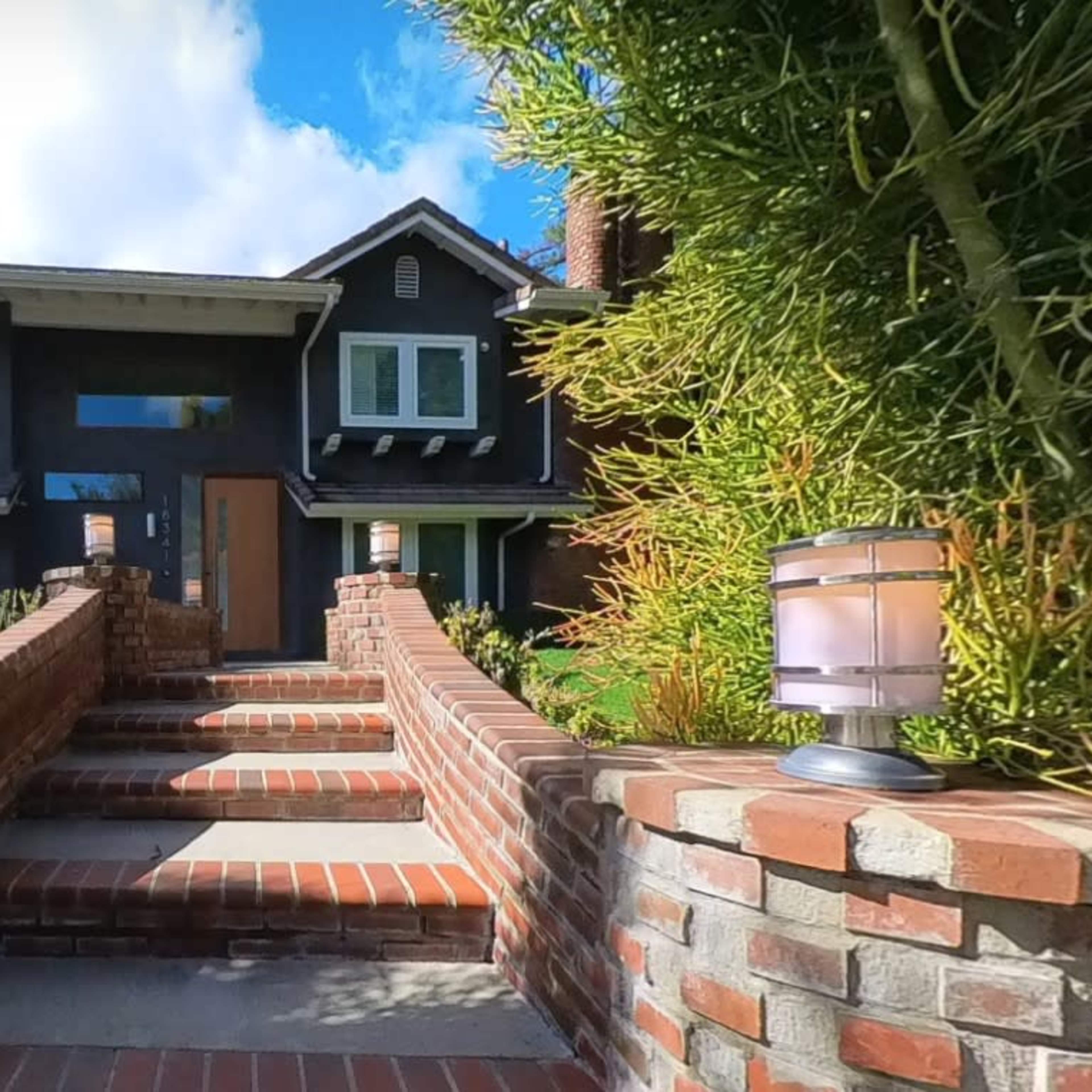 A brick pathway leads to a two-story house with large front windows, flanked by landscaped greenery and outdoor lighting.