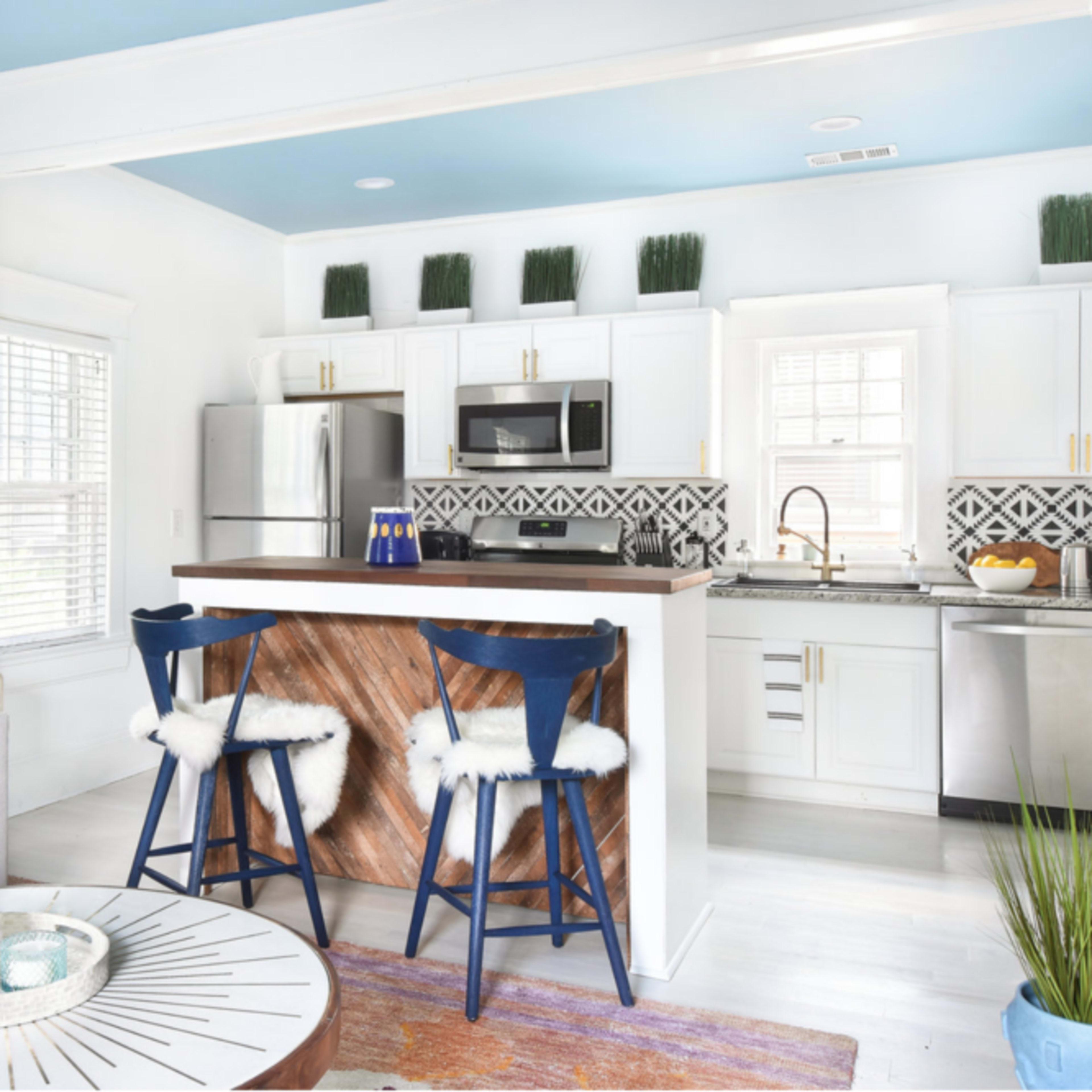 A modern kitchen and living area are connected, featuring a gray sofa, a round coffee table, and a kitchen island with blue stools.