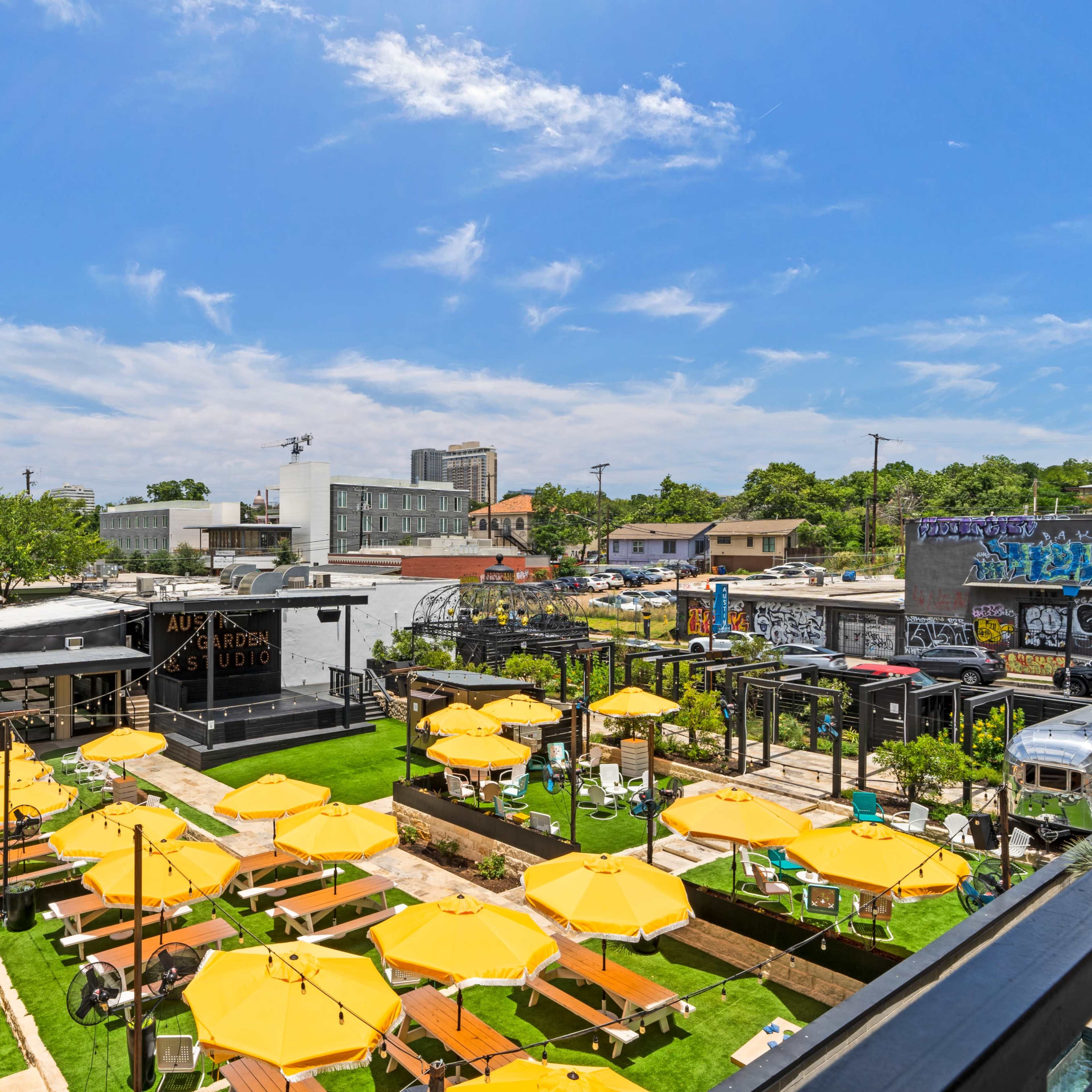 A vibrant outdoor space with yellow umbrellas over tables and a backdrop of buildings and trees under a blue sky.