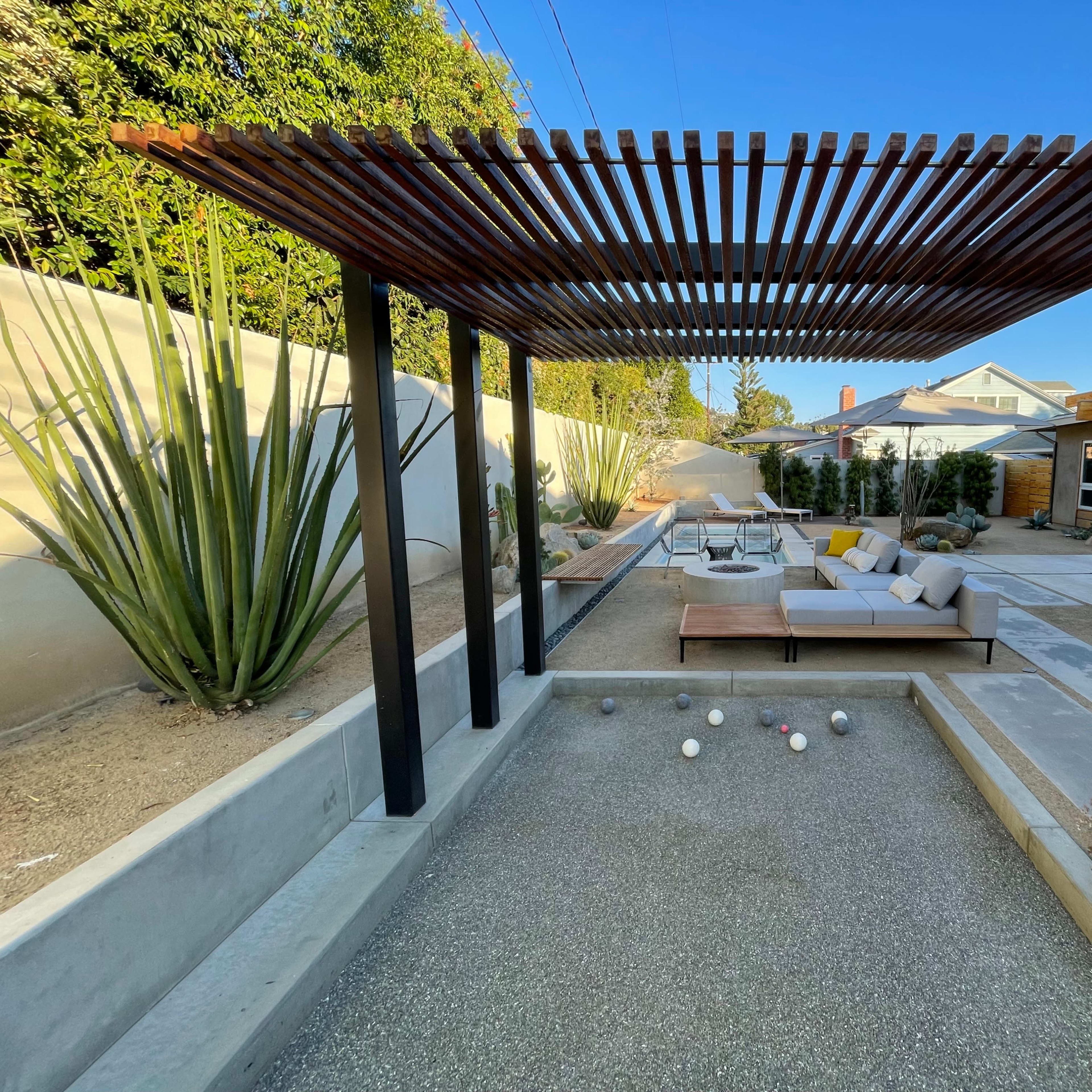 A modern outdoor space featuring a shaded seating area under a wooden trellis, with a gravel bocce ball court in the foreground and various desert plants surrounding the scene.