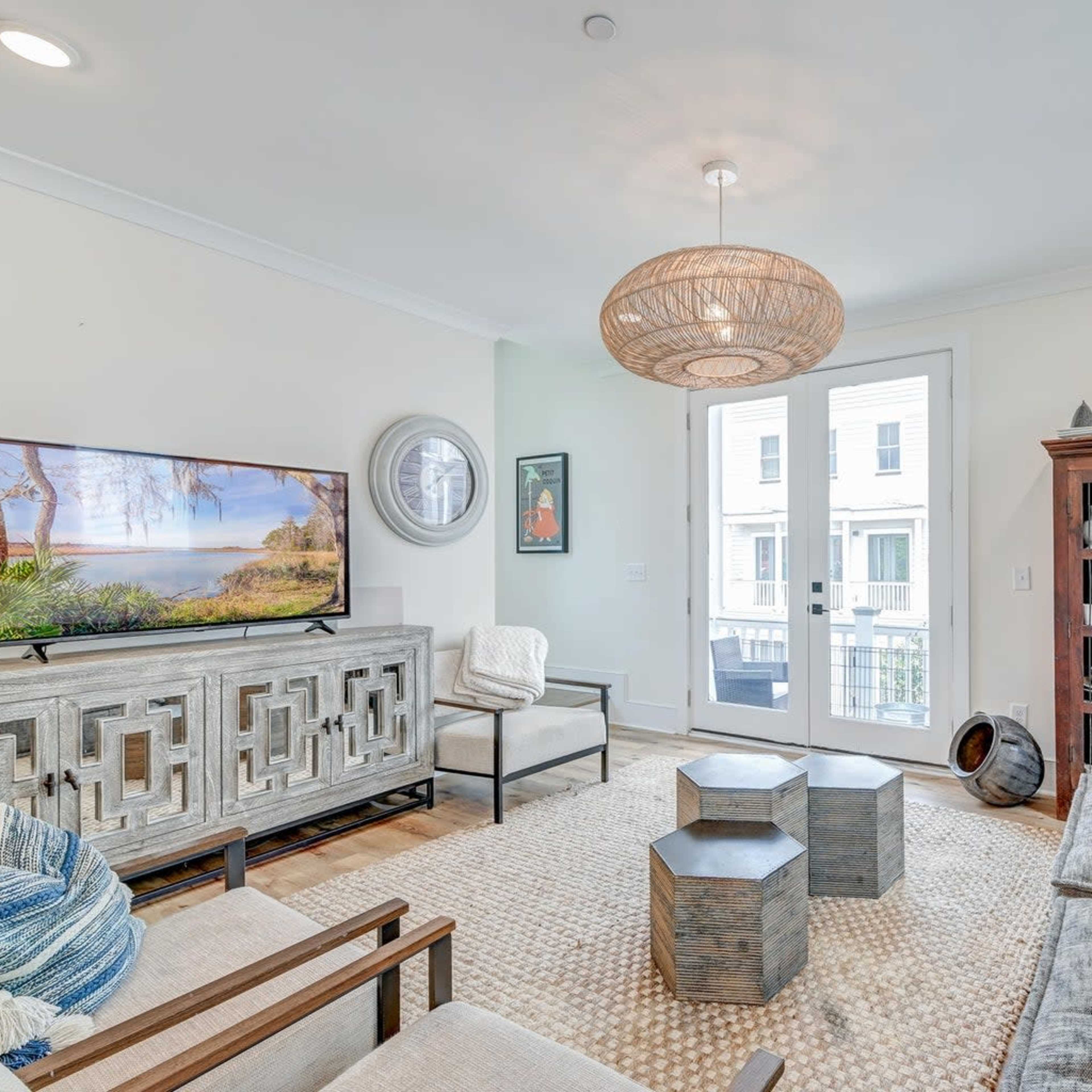 A modern living room with a television, a set of wooden furniture, a woven light fixture, and large windows leading to a balcony.