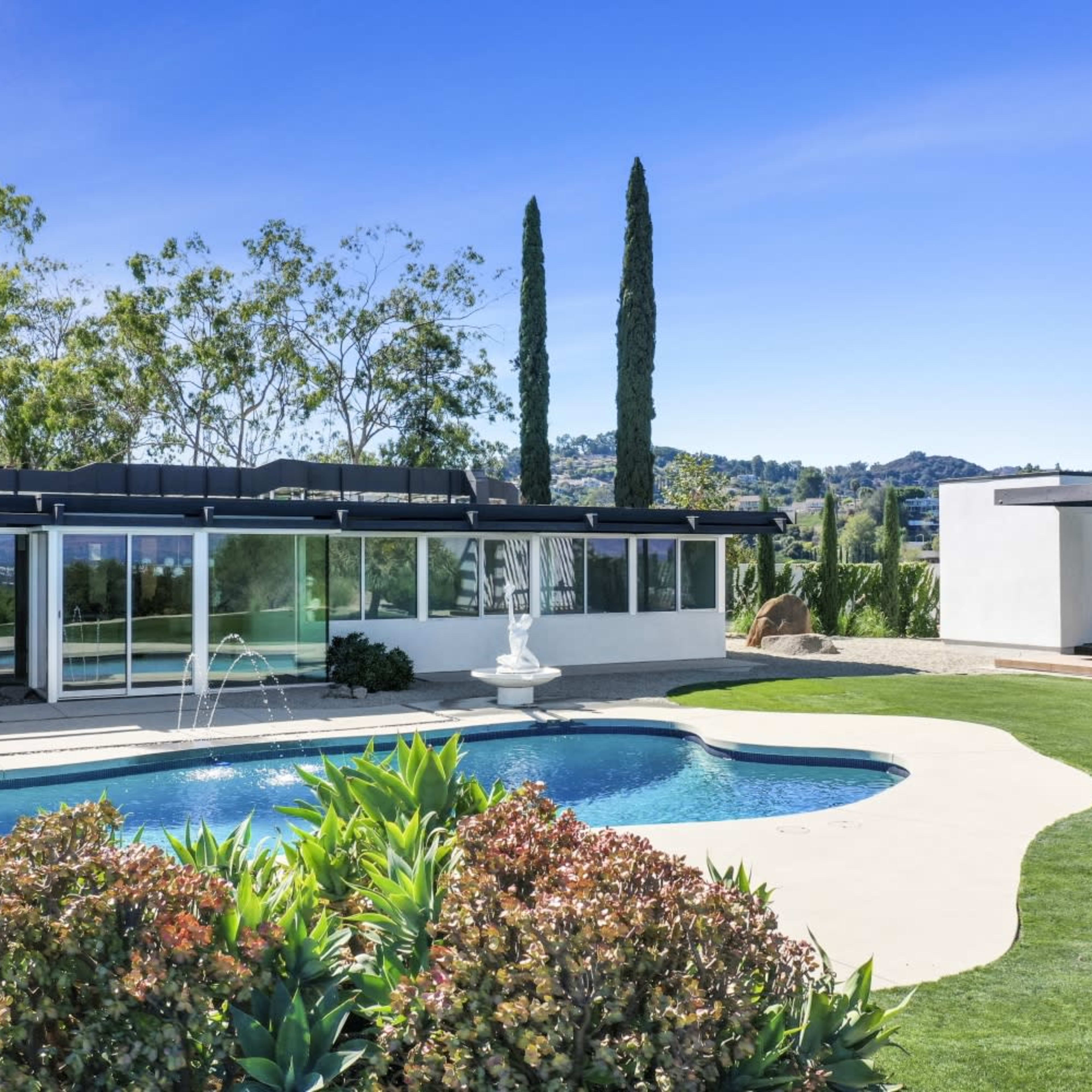 A modern home with large windows overlooks a swimming pool surrounded by landscaping and tall trees.