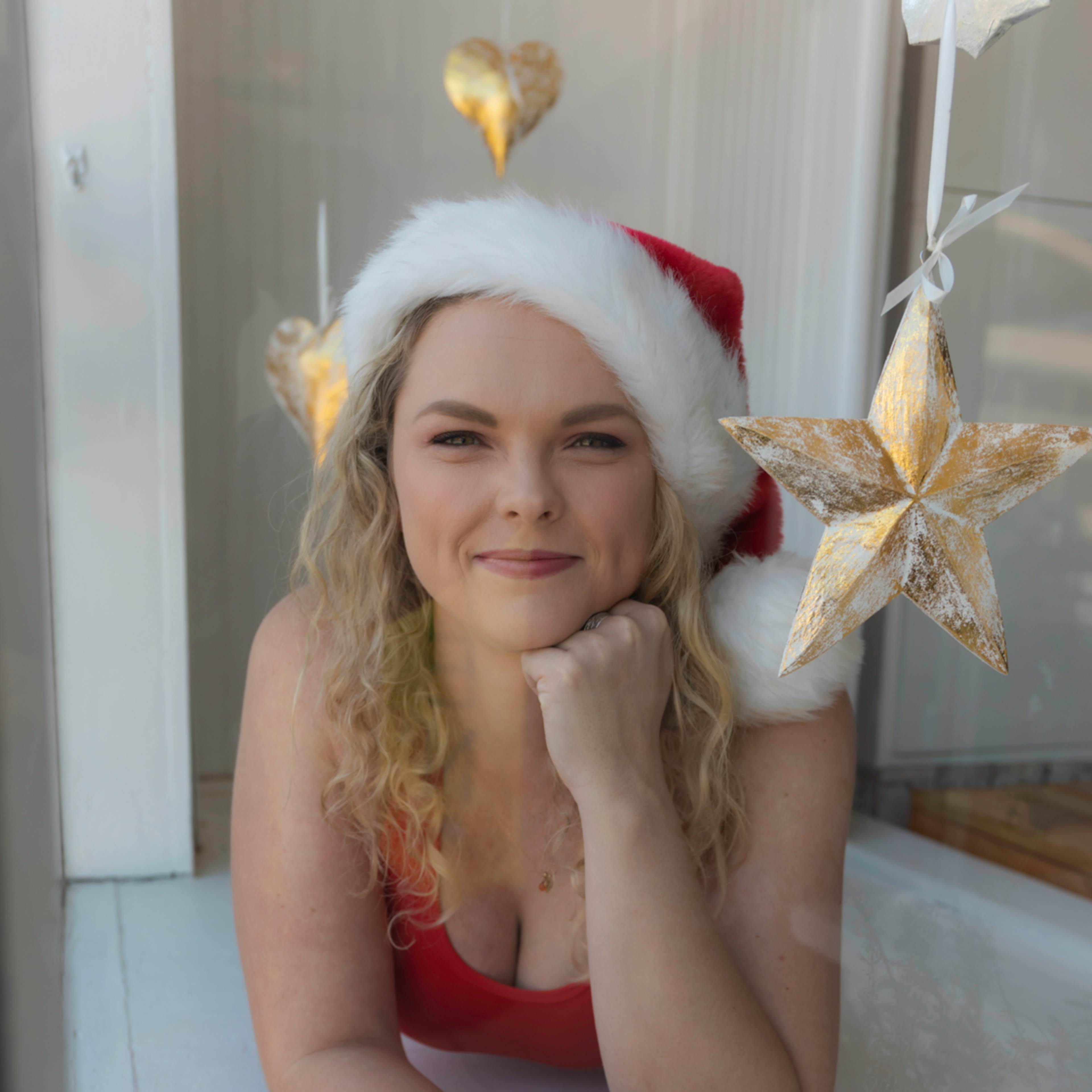 A person wearing a Santa hat and red outfit is resting on a window ledge, posing with her chin on her hand, while gold star decorations hang nearby.