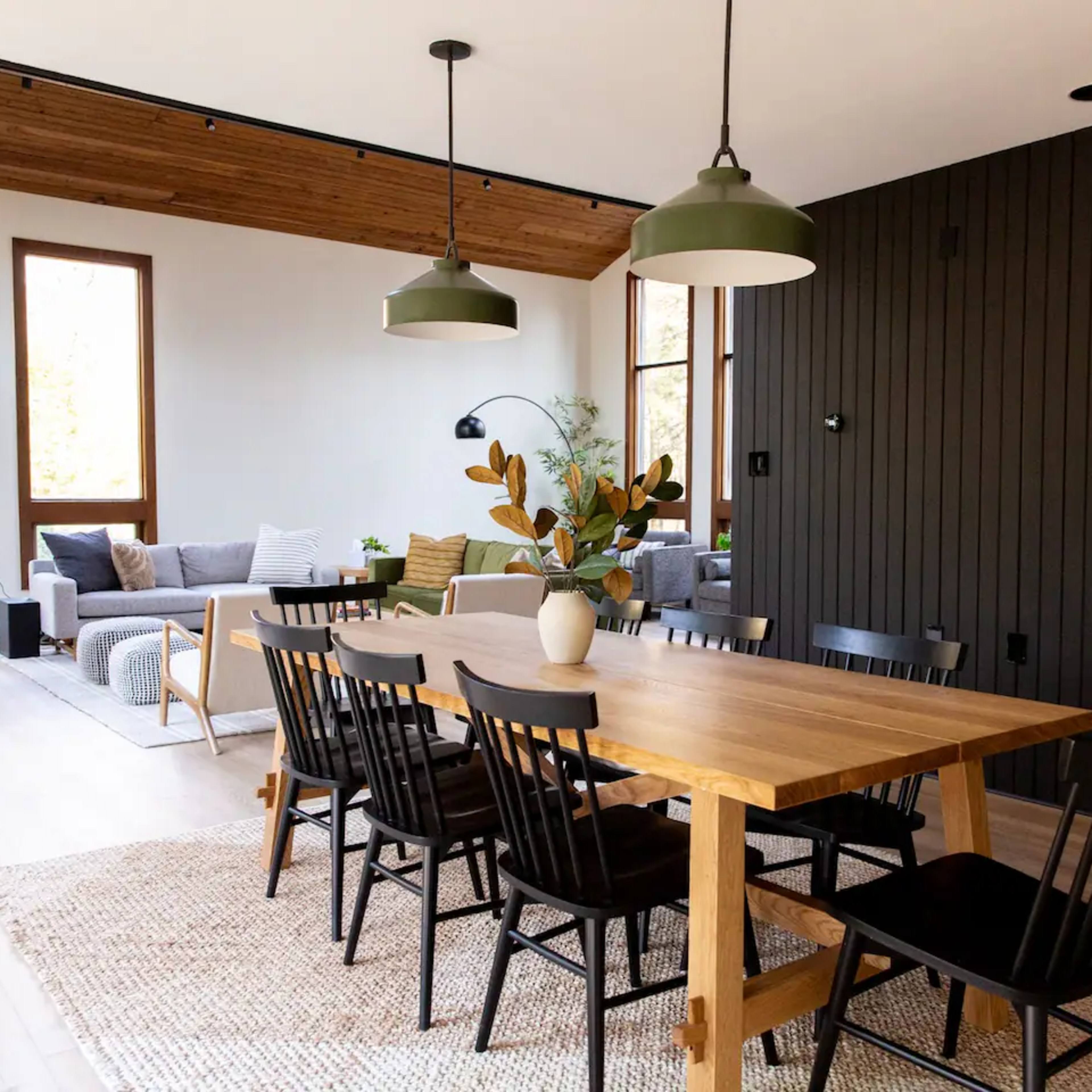 A modern dining area features a large wooden table surrounded by black chairs, with a cozy seating area and large windows in the background.