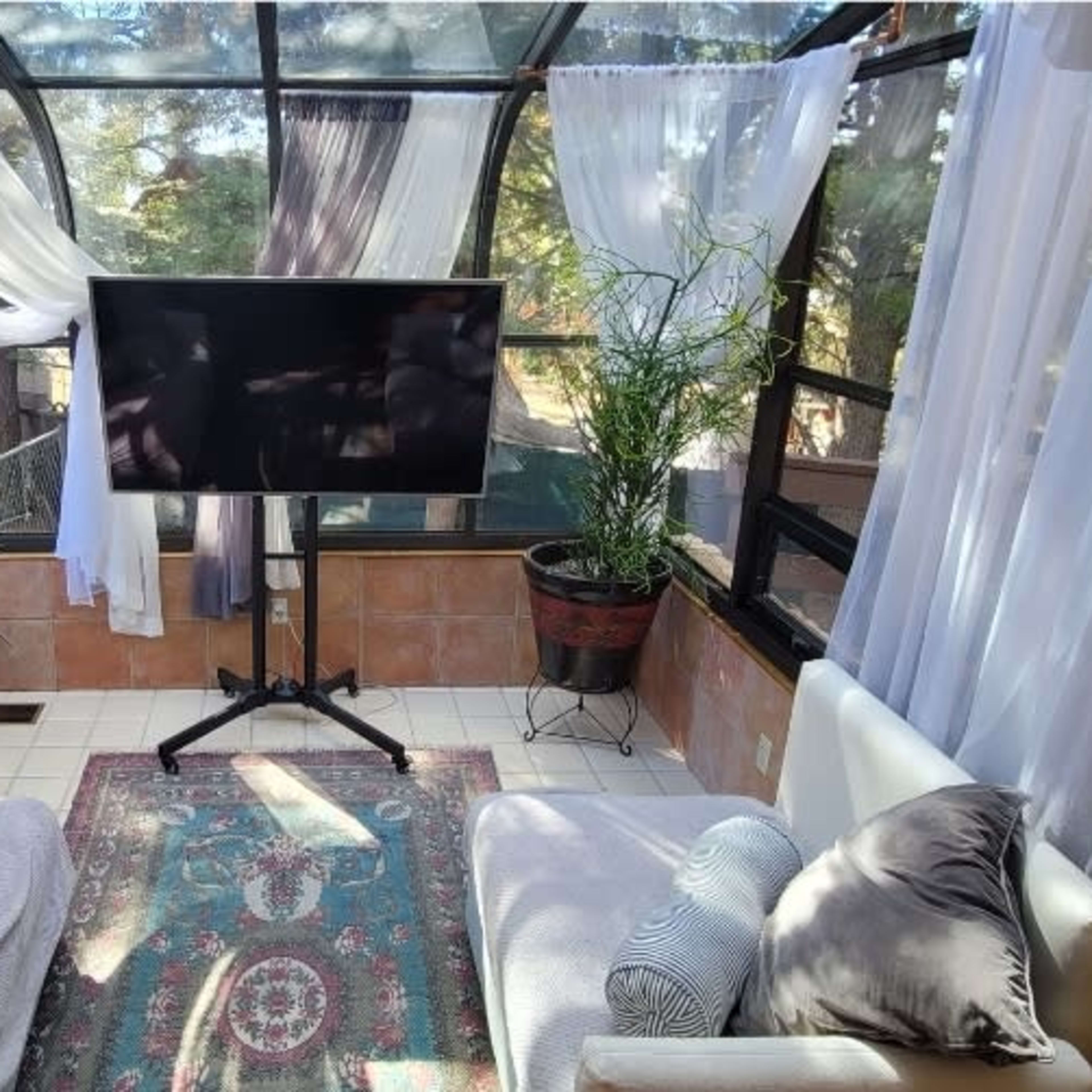 A sunlit sunroom features white curtains, two couches, a television on a stand, and potted plants.