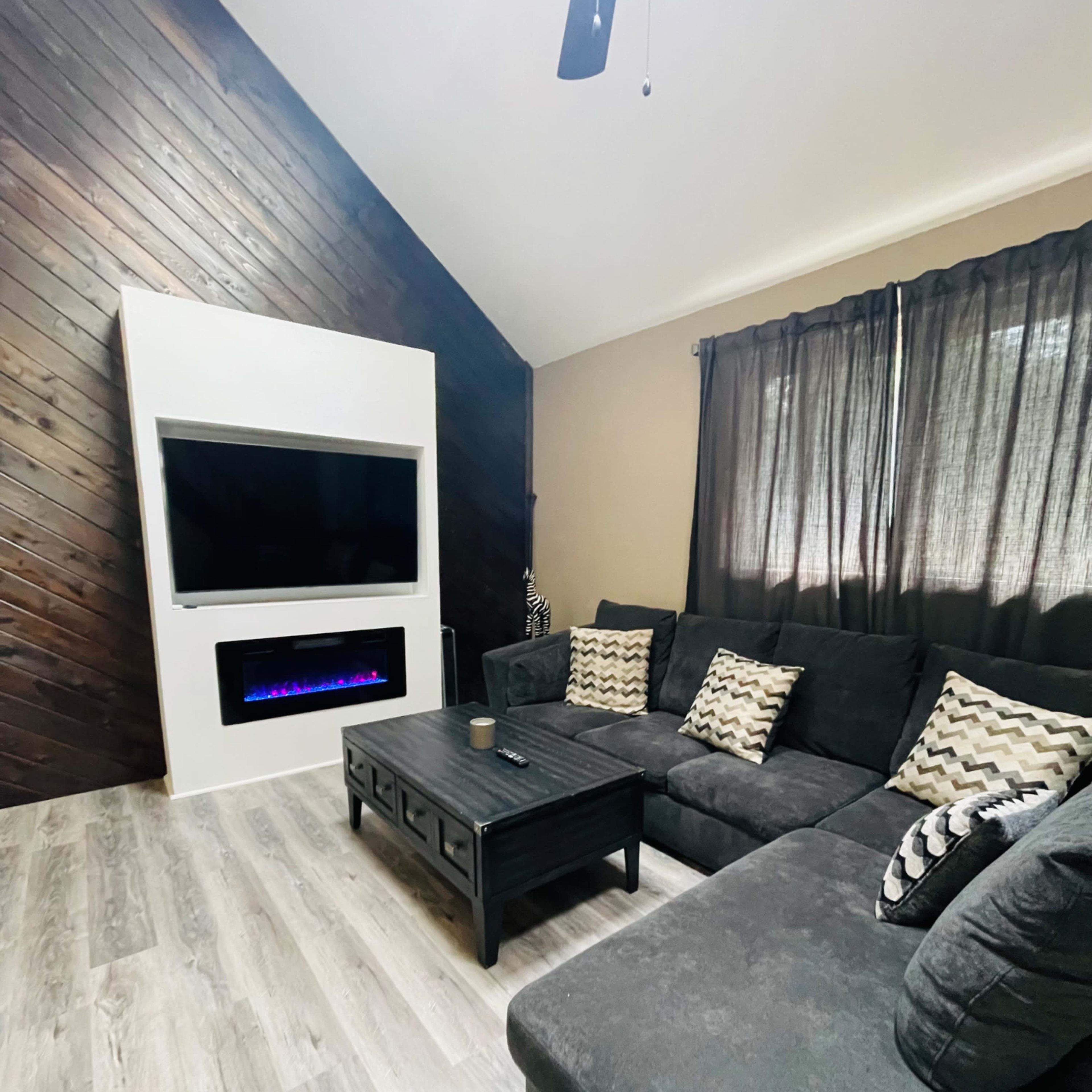 A modern living room with a sectional sofa, a coffee table, an electric fireplace beneath a mounted television, and dark curtains on the windows.
