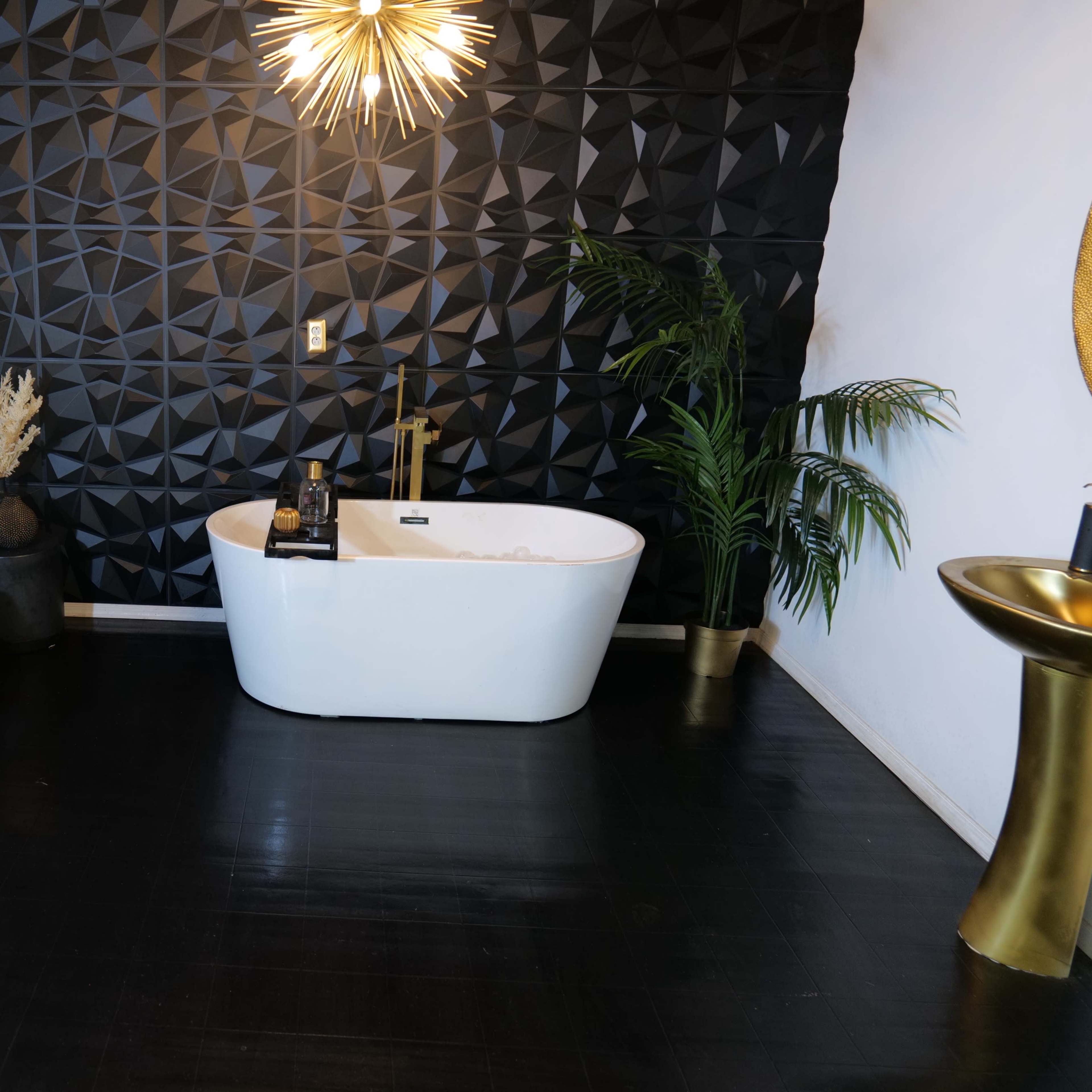 A modern bathroom with a freestanding white bathtub, a geometric black accent wall, a potted plant, and decorative gold elements.