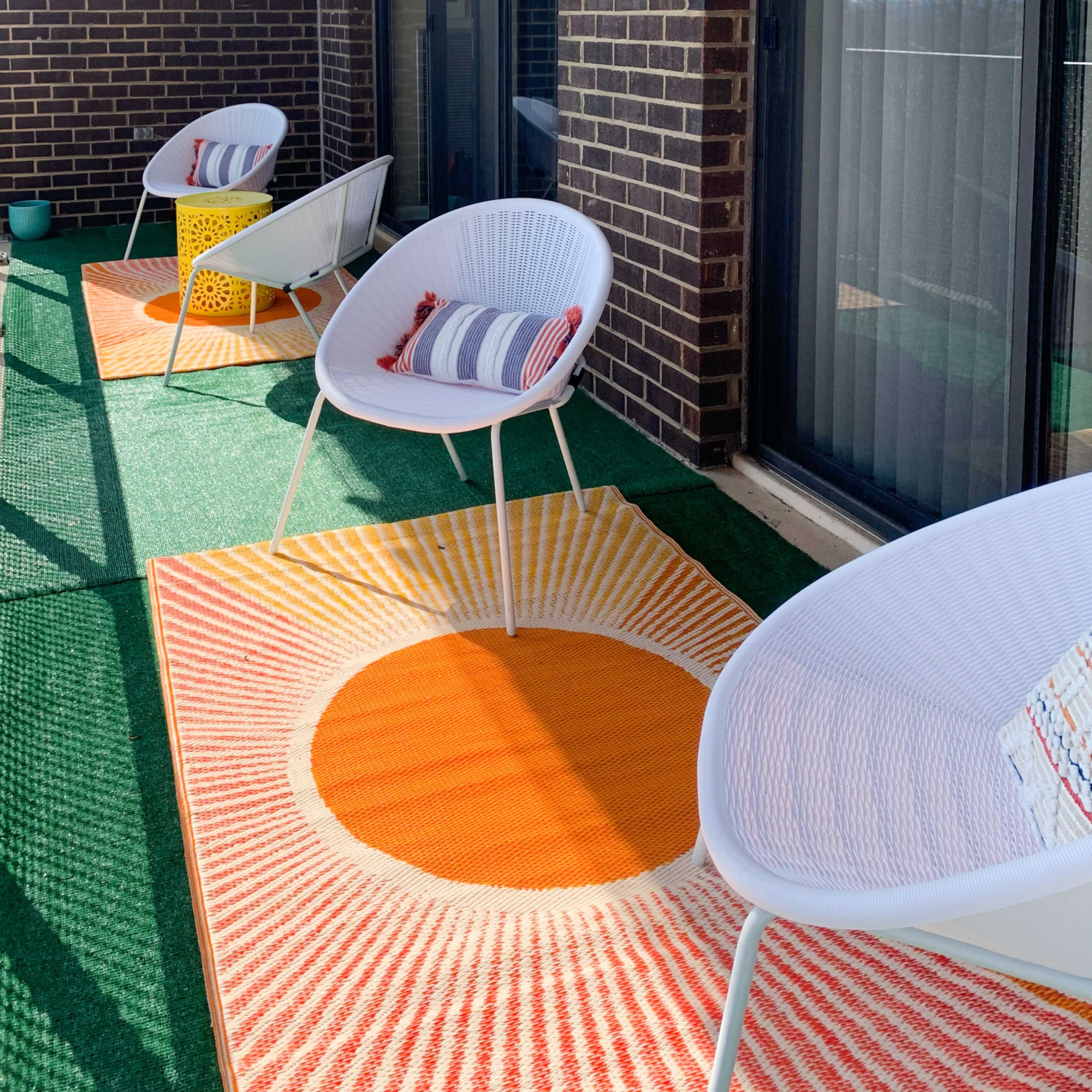 A balcony with four white chairs arranged on a green carpet, featuring an orange and yellow sun-patterned rug.