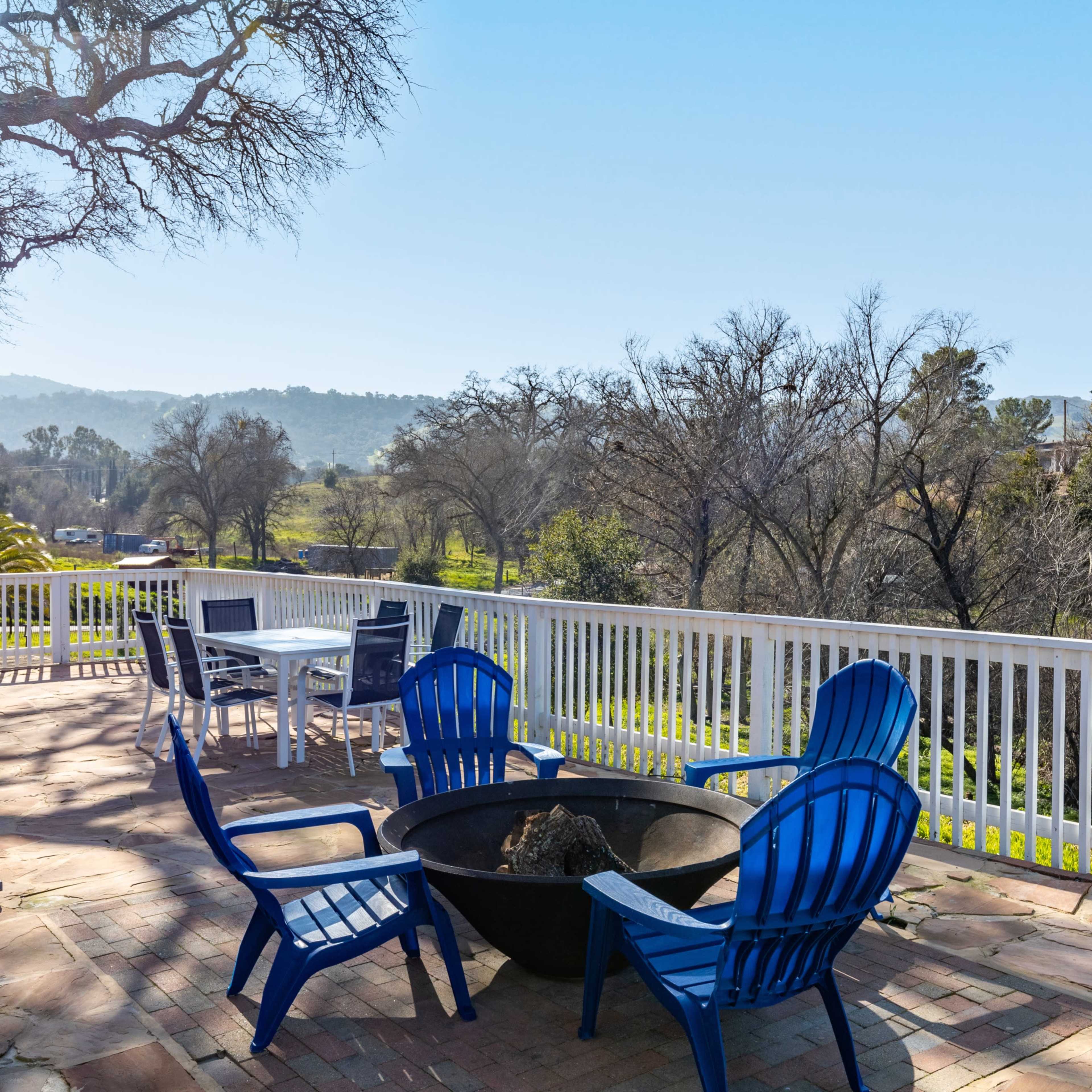 A patio features blue chairs surrounding a fire pit, with a table set for dining and a scenic view of trees and hills in the background.