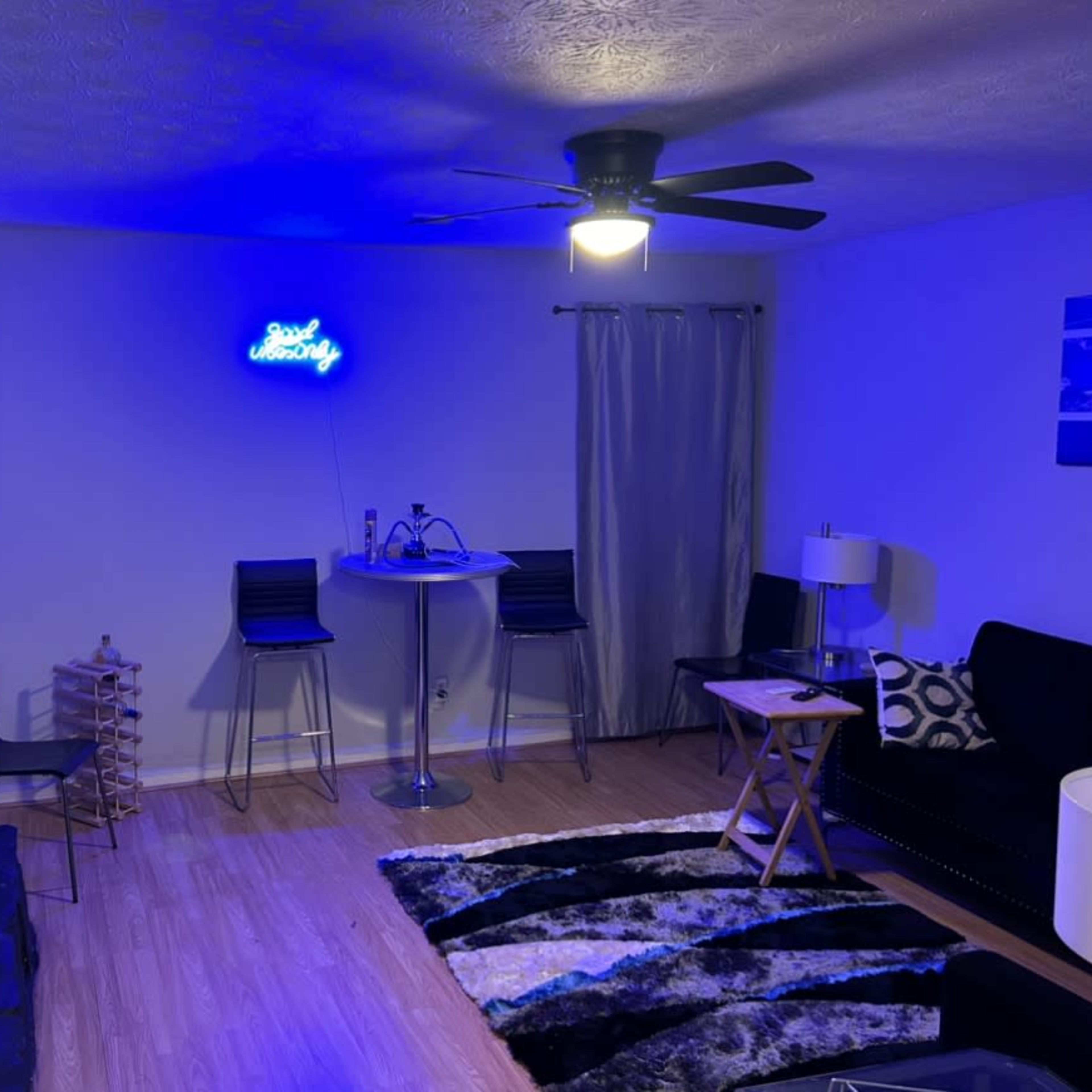 A modern living room features a black couch, a round bar table with stools, a wall-mounted television, and a neon sign against a light-colored wall.