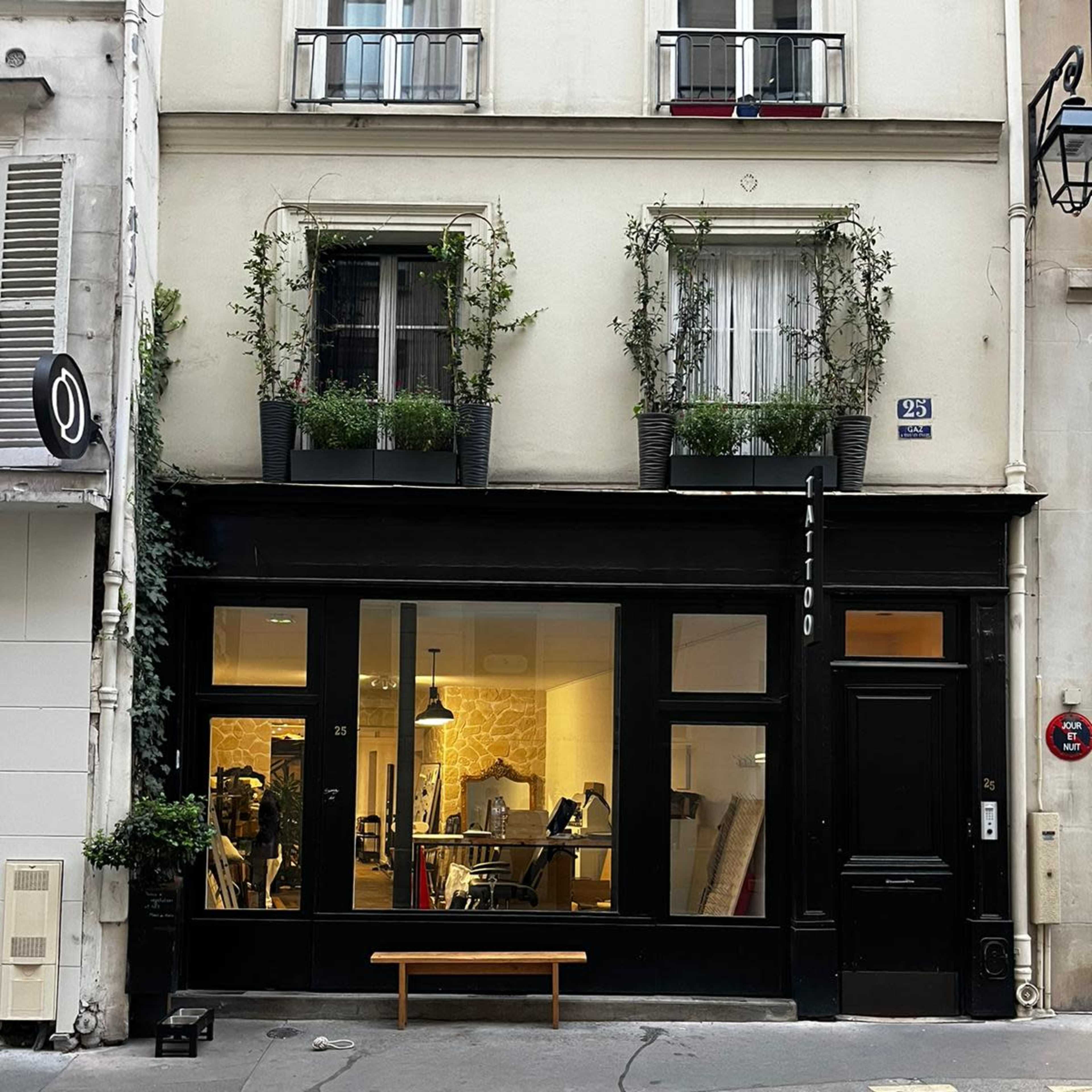 A black storefront with large windows displaying a tattoo studio, flanked by plants in window boxes, is situated on a quiet street in Paris.