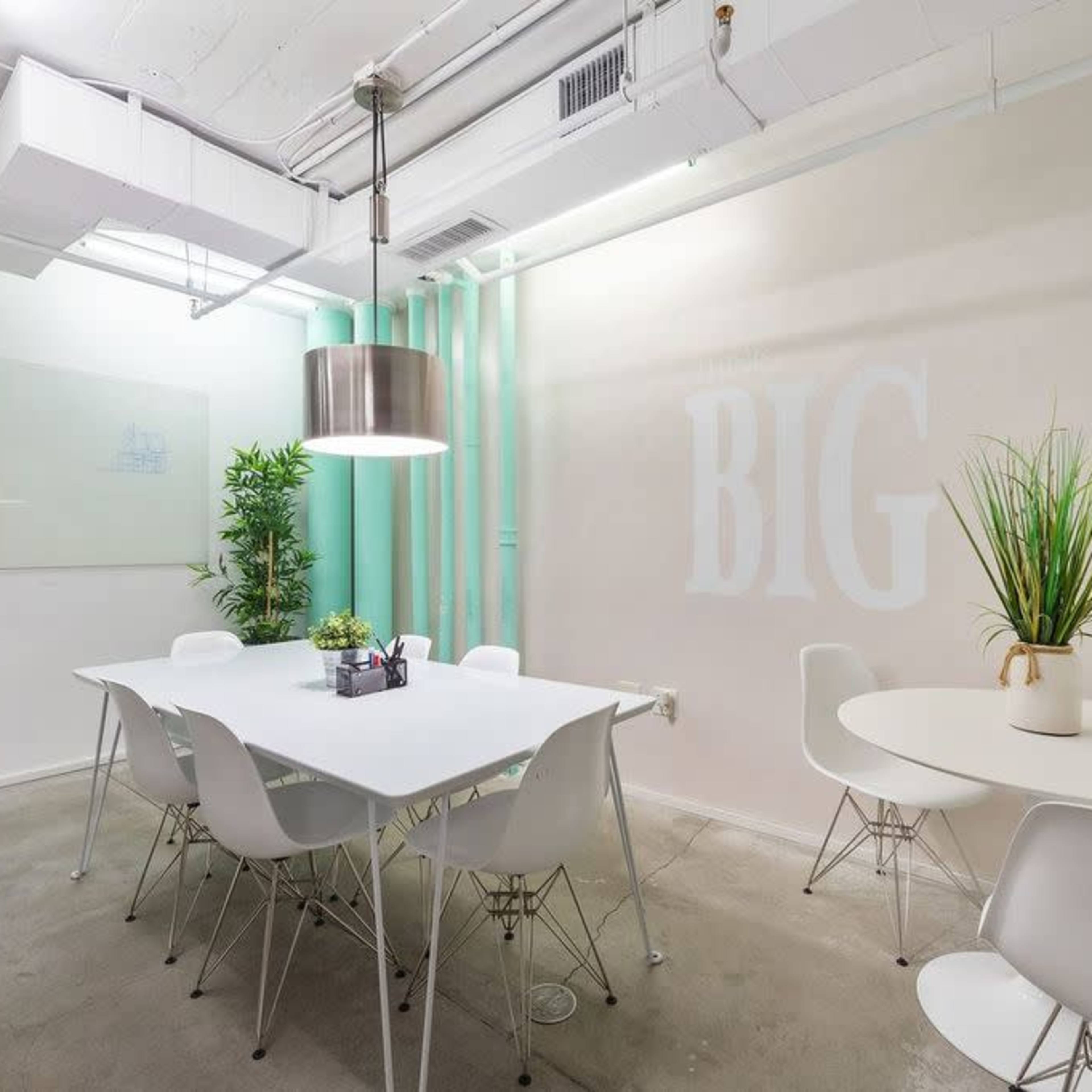 A well-lit meeting room features a white table surrounded by chairs, along with plants and a large pendant light.