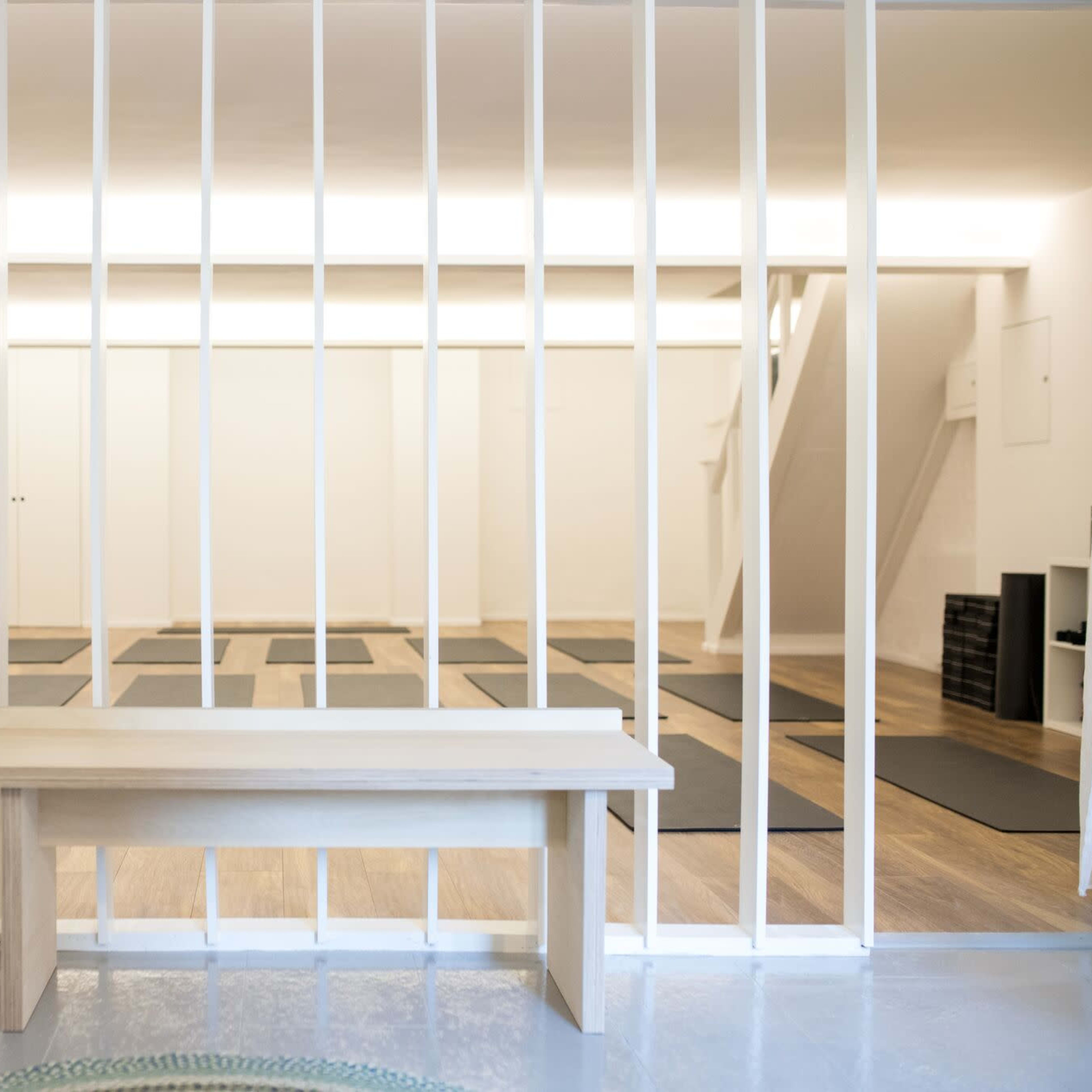 A minimalist yoga studio with several mats arranged on the floor, framed by white wooden slats and featuring a potted plant in the foreground.