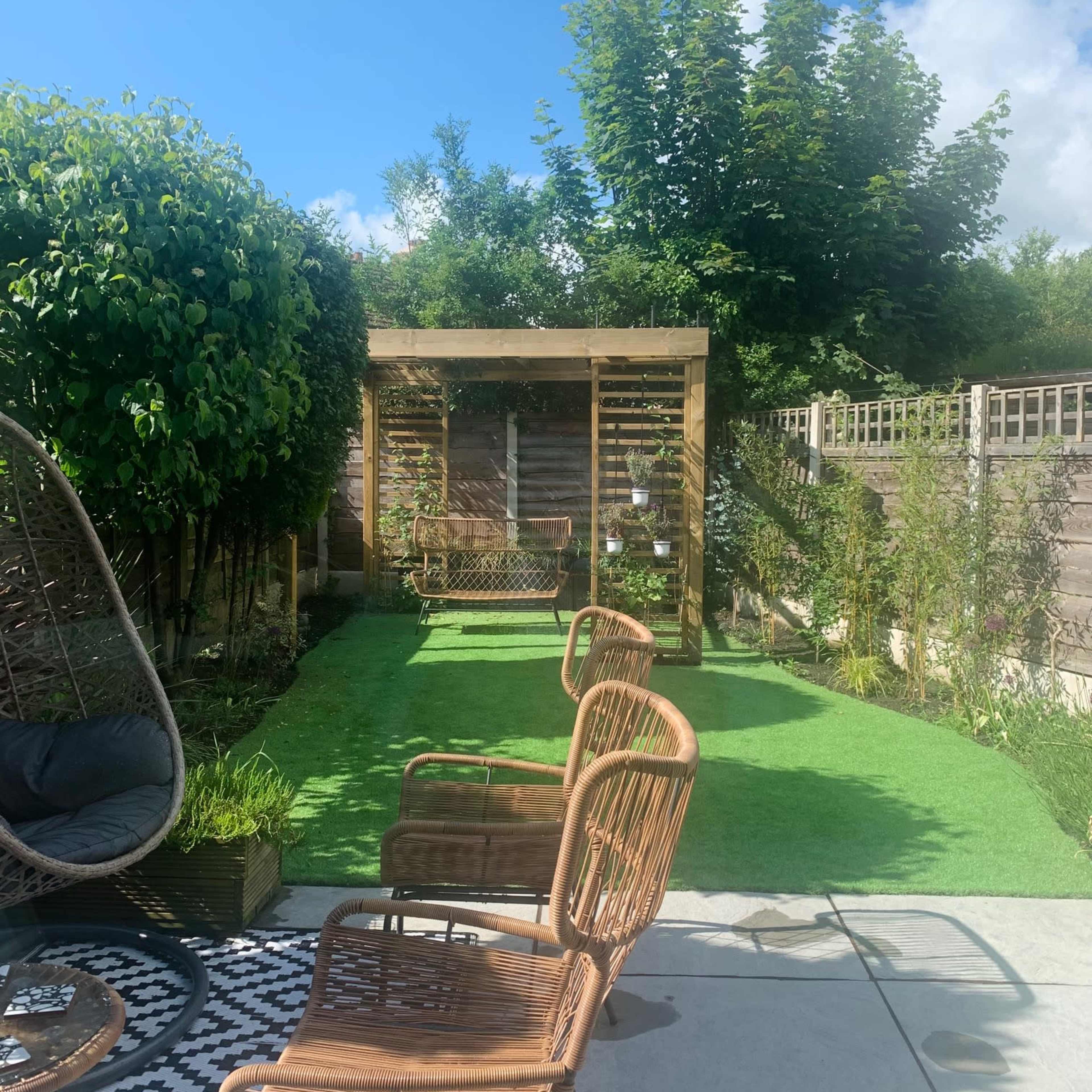A well-maintained garden with artificial grass, featuring wooden benches and a decorative arbor in the background, surrounded by greenery.