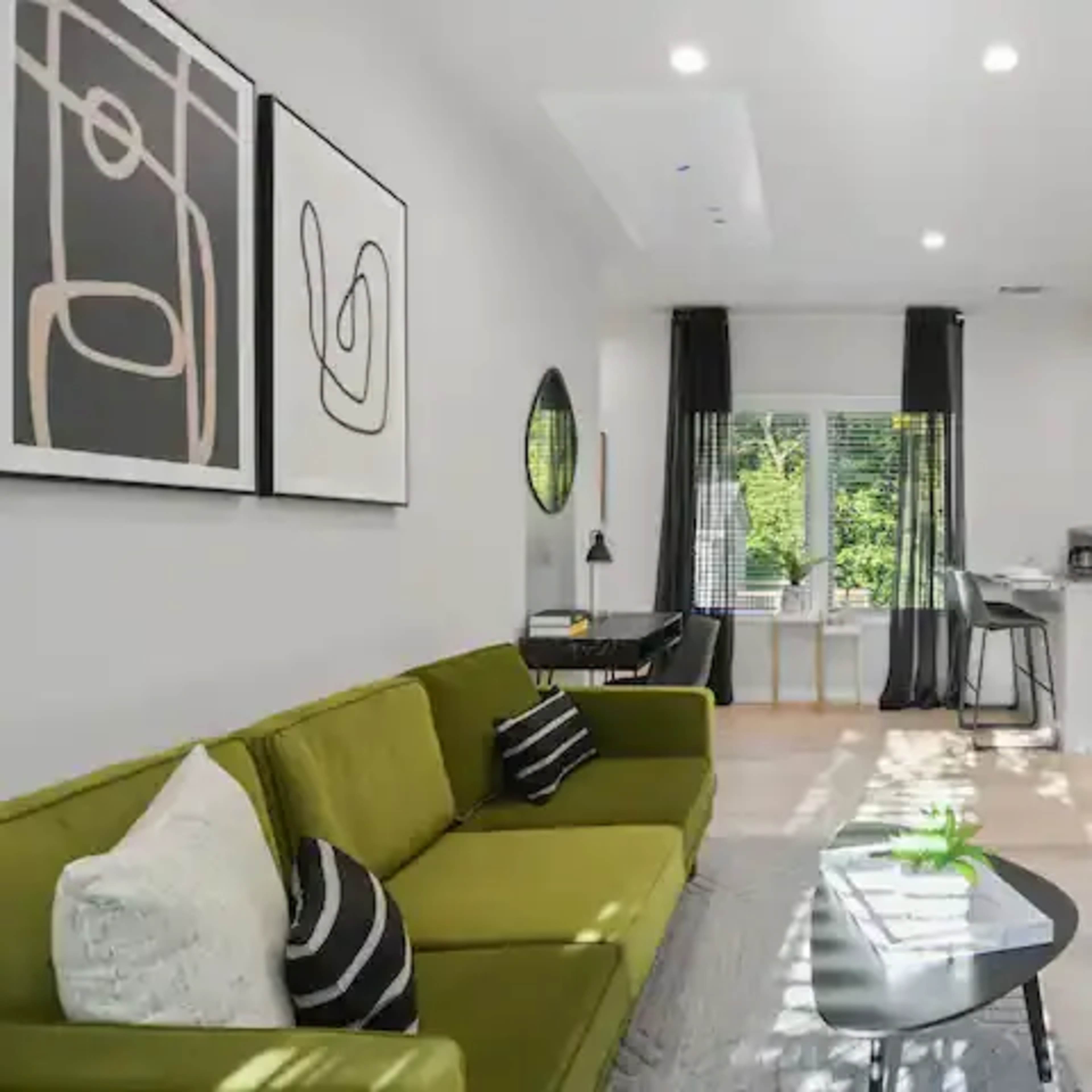 A modern living room features a green sofa, abstract wall art, and a large flat-screen TV, with sunlight streaming through the windows.