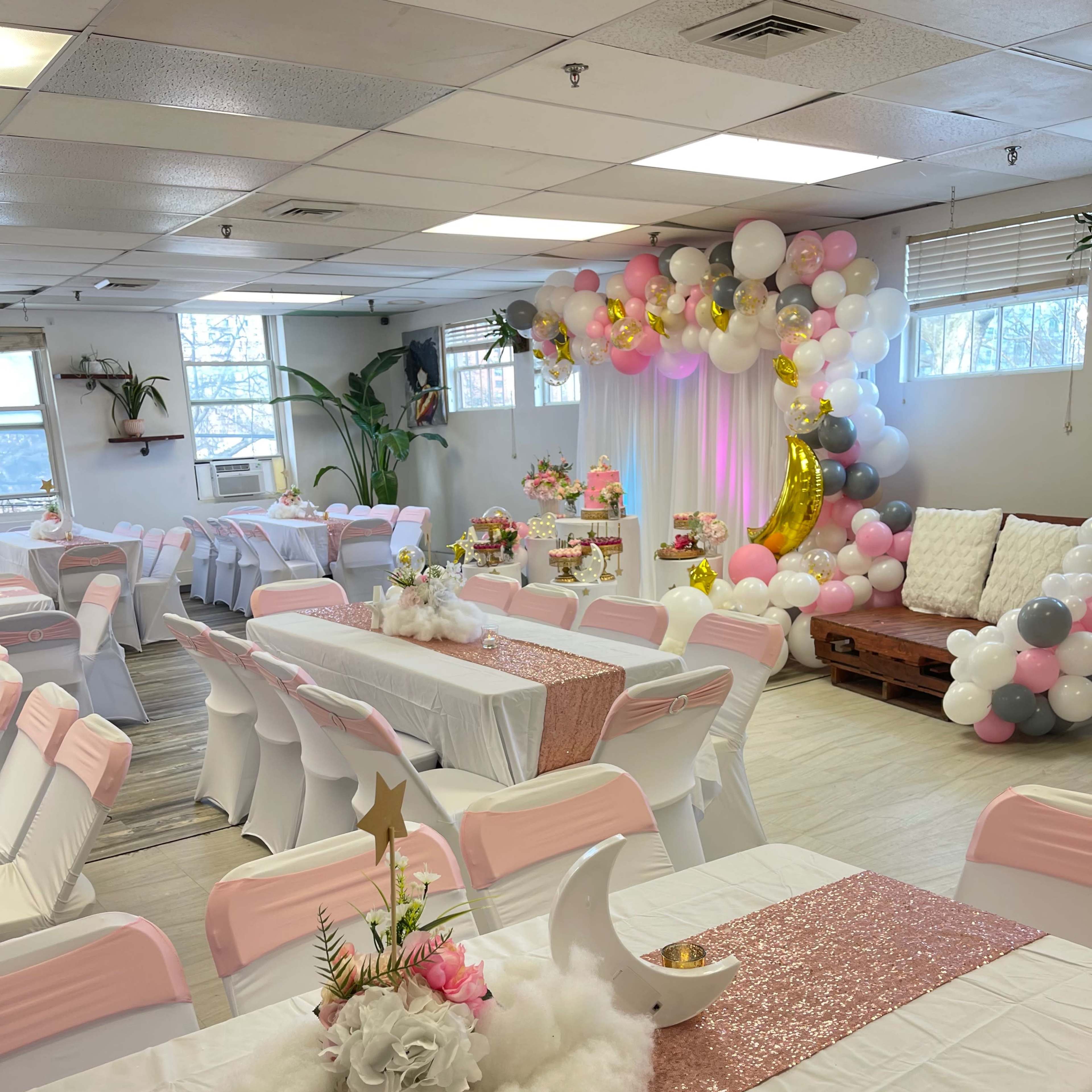 A decorated event space with tables covered in white tablecloths and pink accents, adorned with floral centerpieces and a colorful balloon arch in the background.