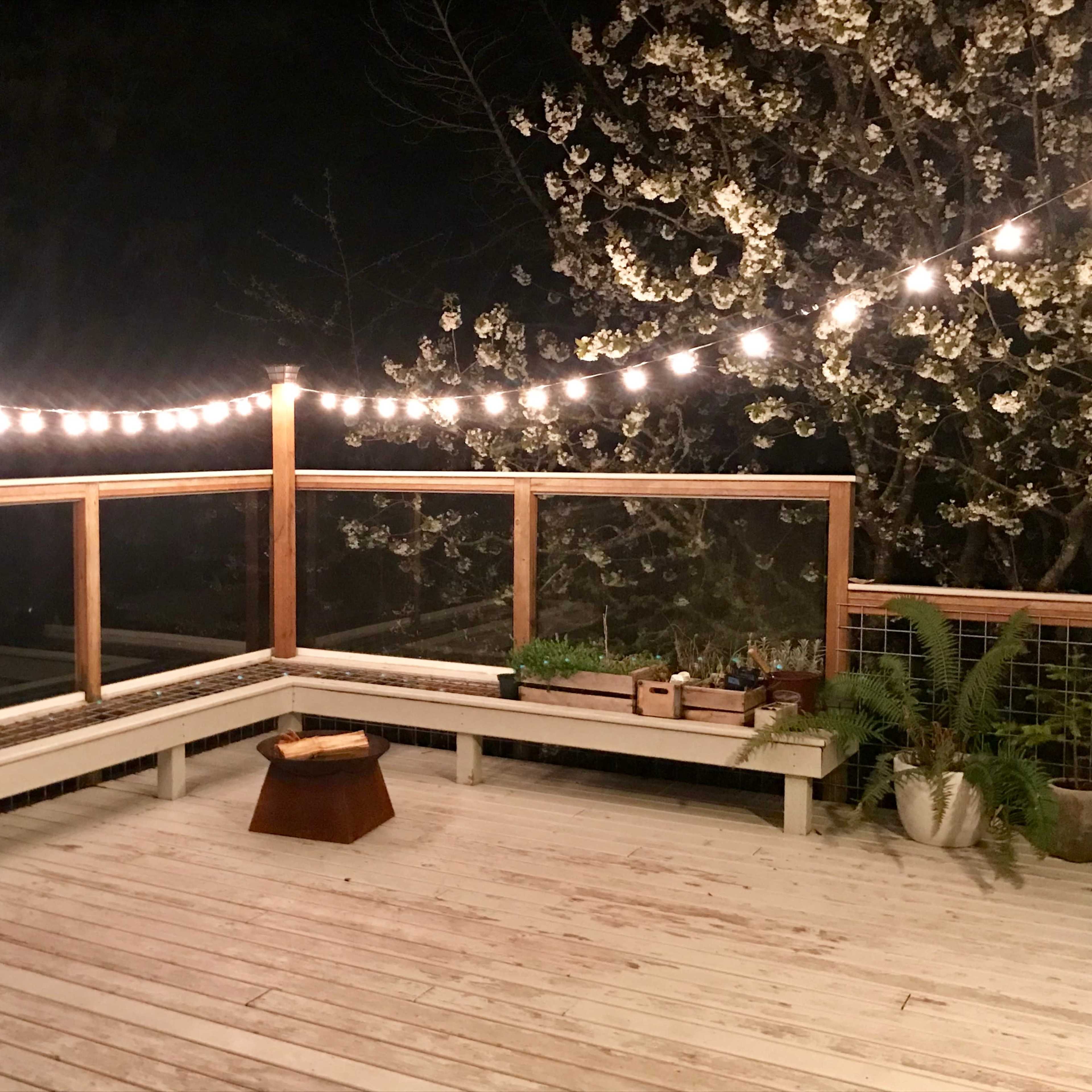 A wooden deck features a seating area surrounded by potted plants and illuminated by string lights at night.
