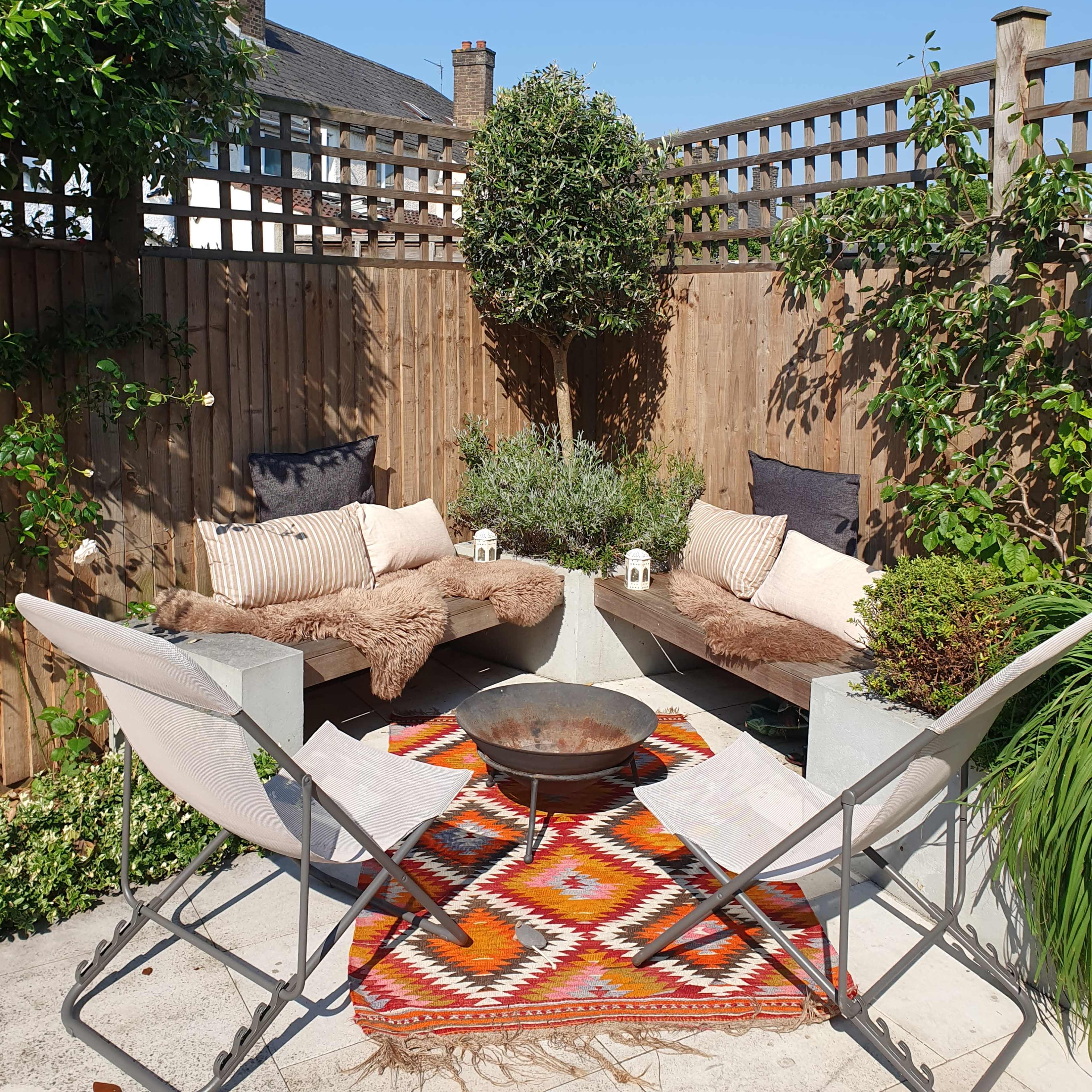 A cozy outdoor seating area features two benches and two chairs surrounding a fire pit, with a decorative rug in the center and greenery against a wooden fence.