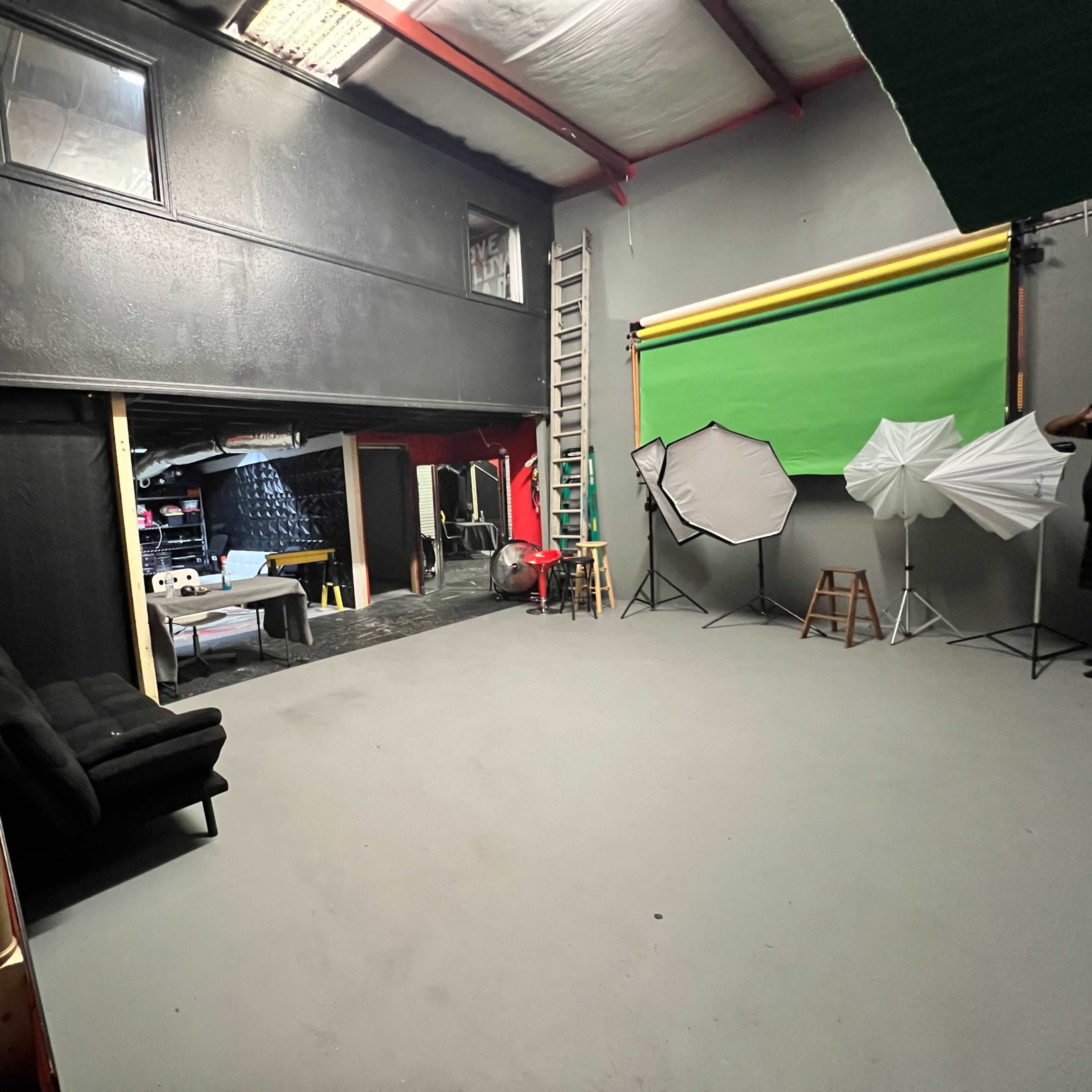 A spacious studio with a gray floor, black walls, a green backdrop, various lighting equipment, and a staircase leading to an upper level.