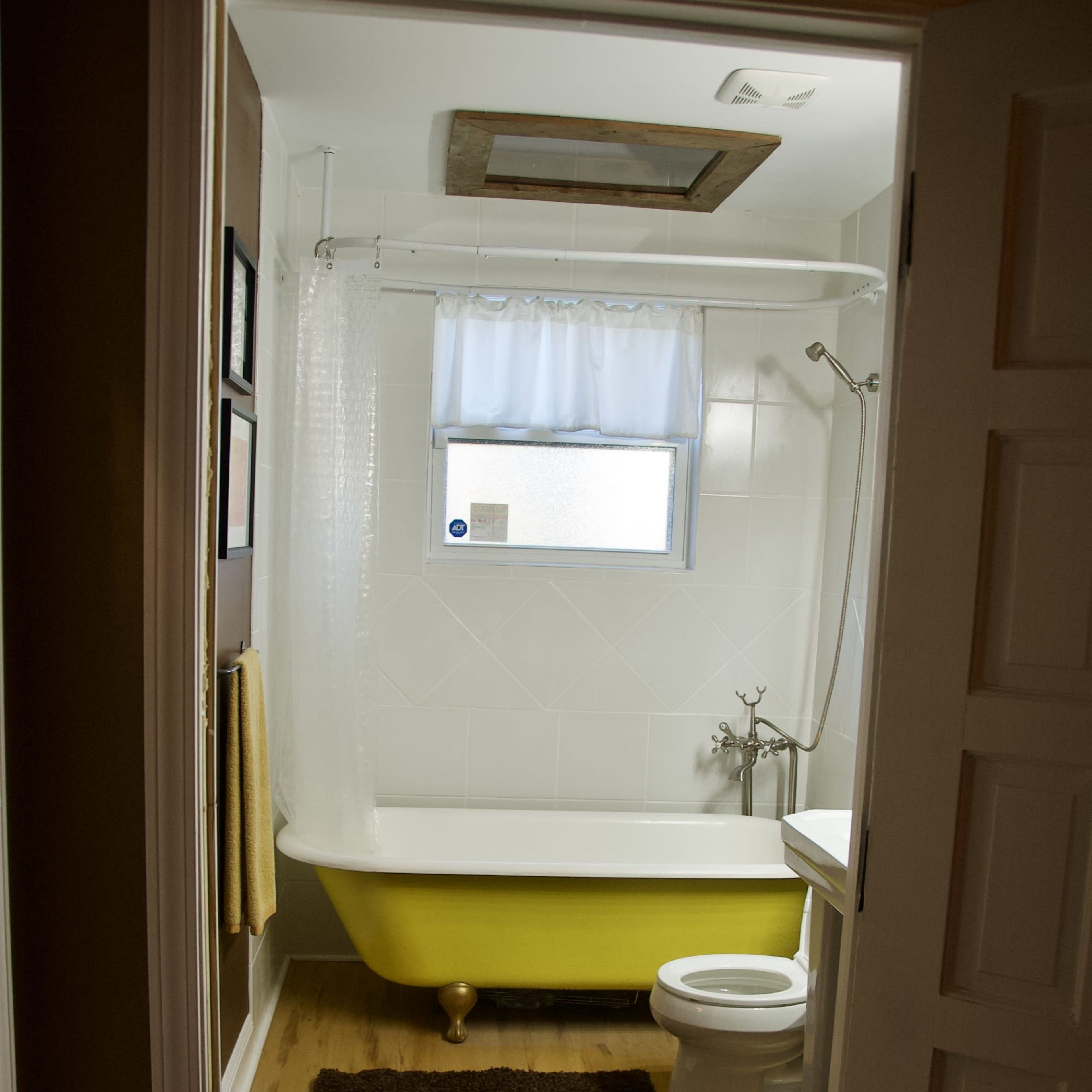 A bathroom featuring a yellow clawfoot bathtub, a toilet, and a small rug on a wooden floor.