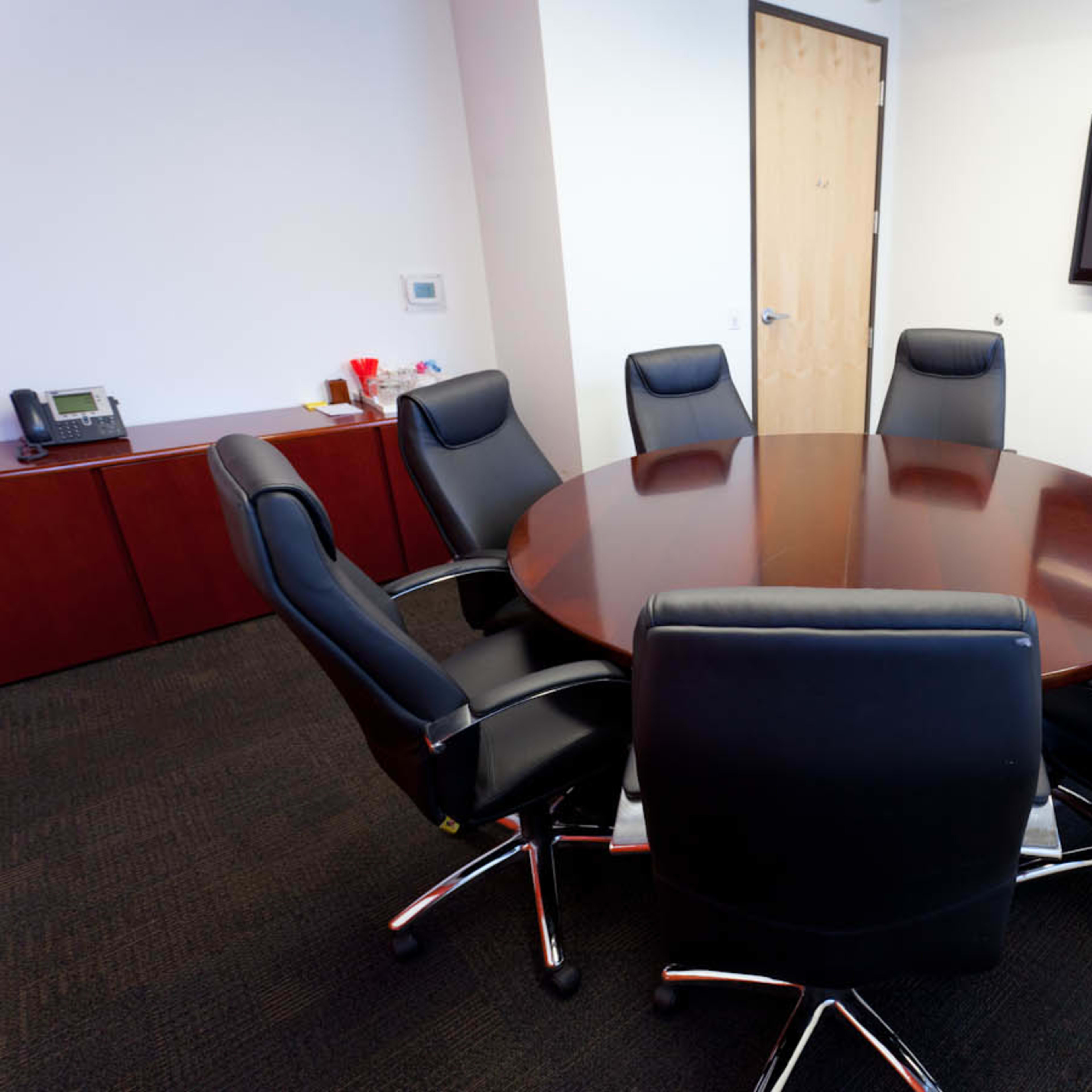 A round wooden conference table surrounded by seven black leather chairs in a modern meeting room.