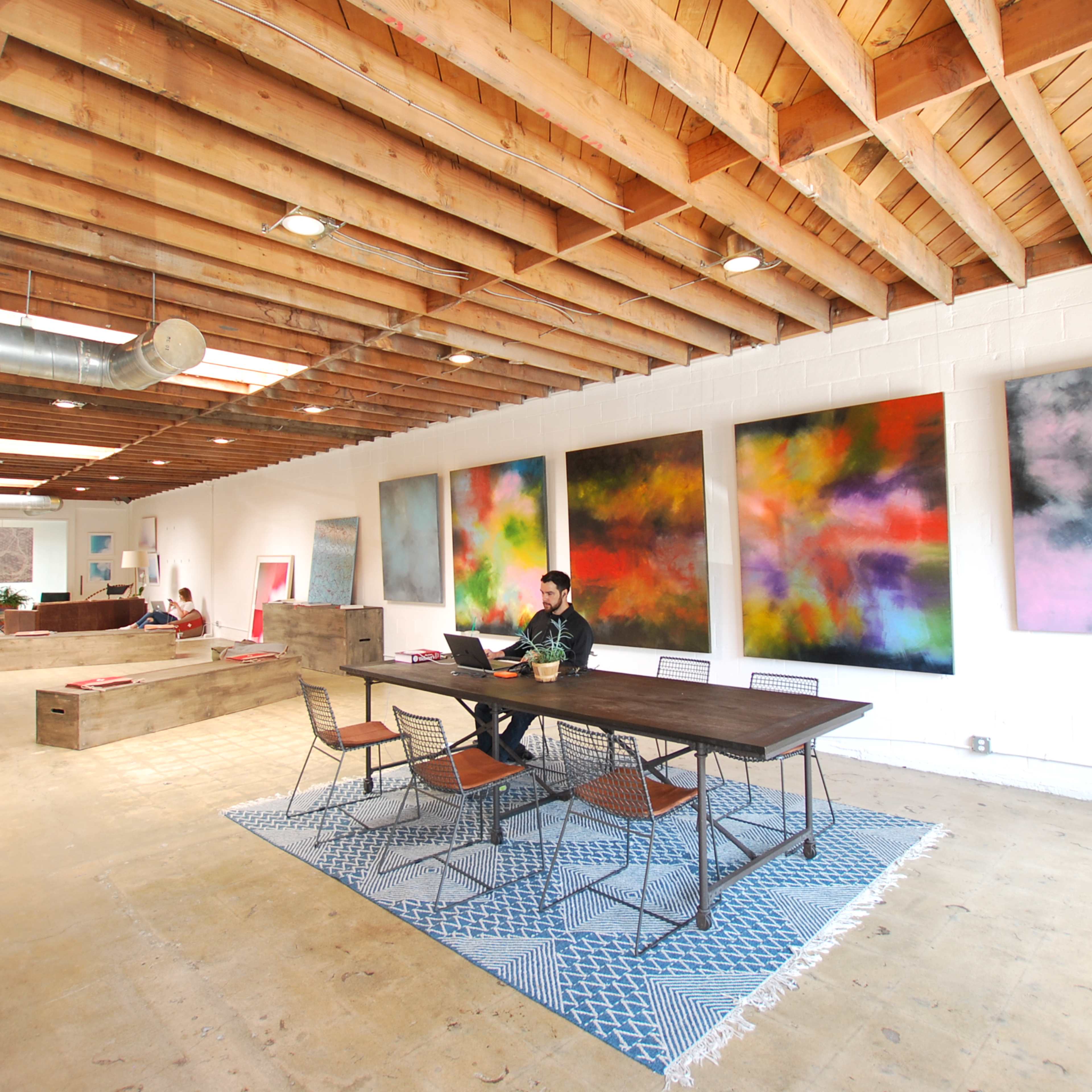 A person works at a table in a spacious room with high wooden ceilings and colorful abstract paintings on the walls.