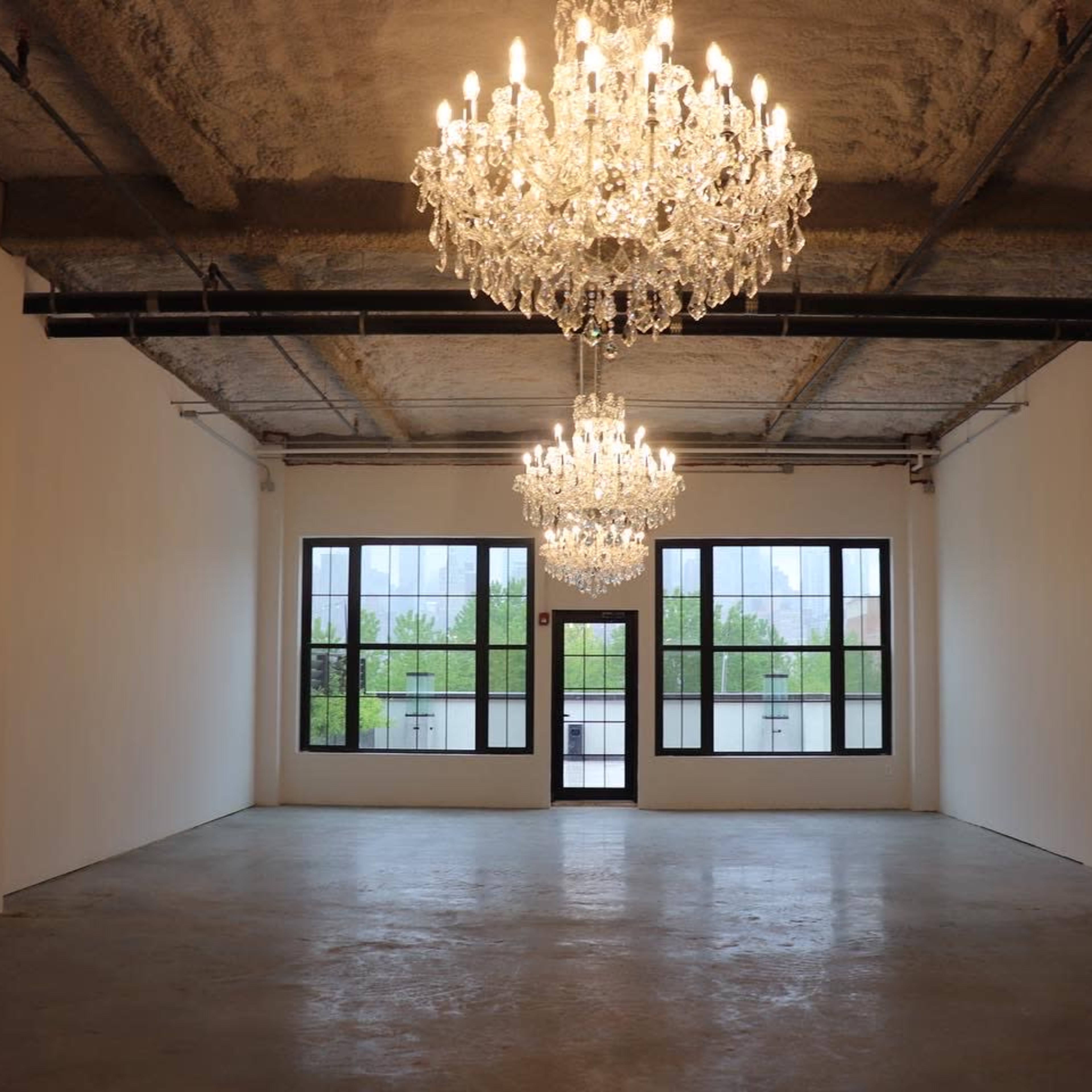 A spacious room with two large chandeliers hanging from the ceiling and four windows providing natural light.