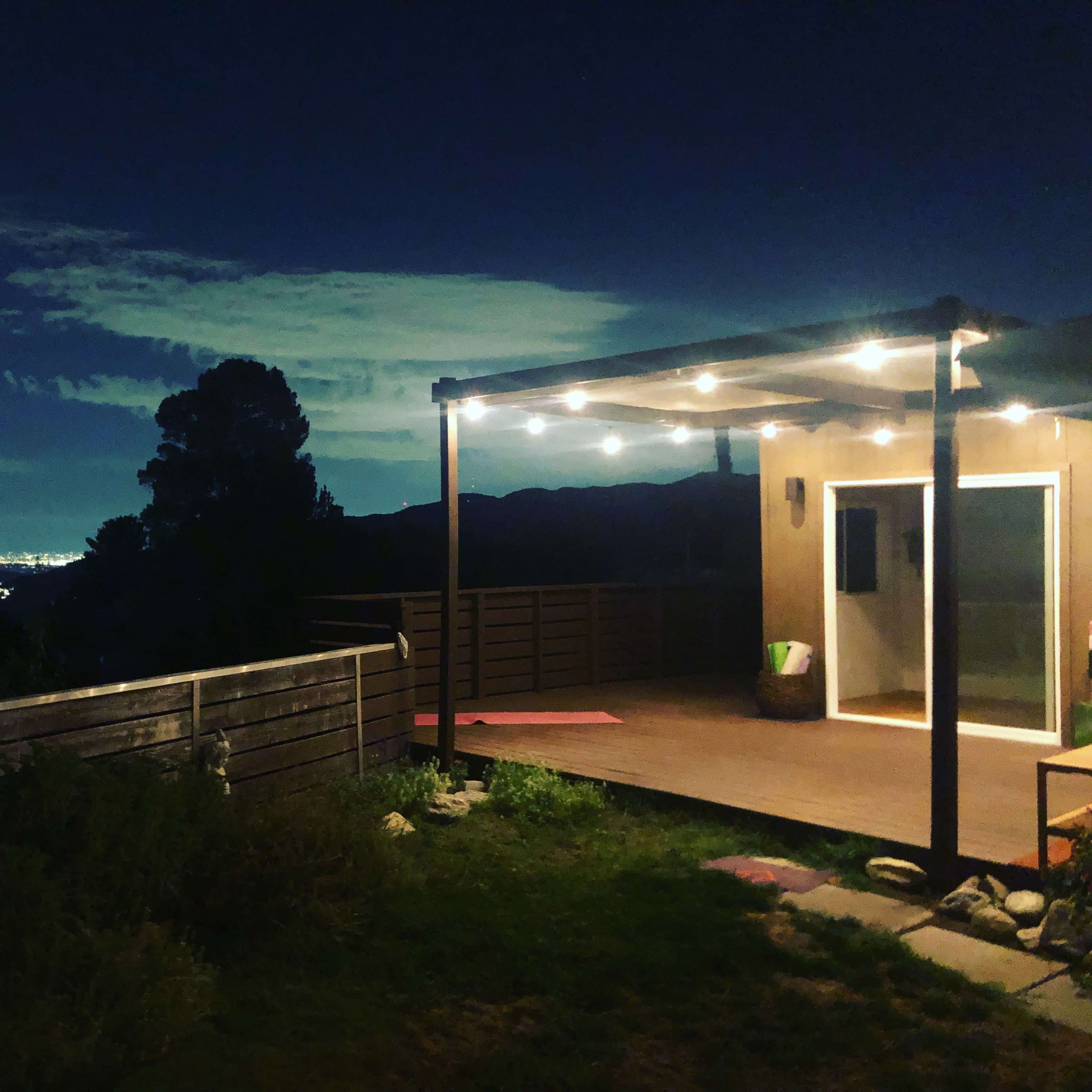 A deck with string lights overlooks a city skyline at night, surrounded by trees and plants.