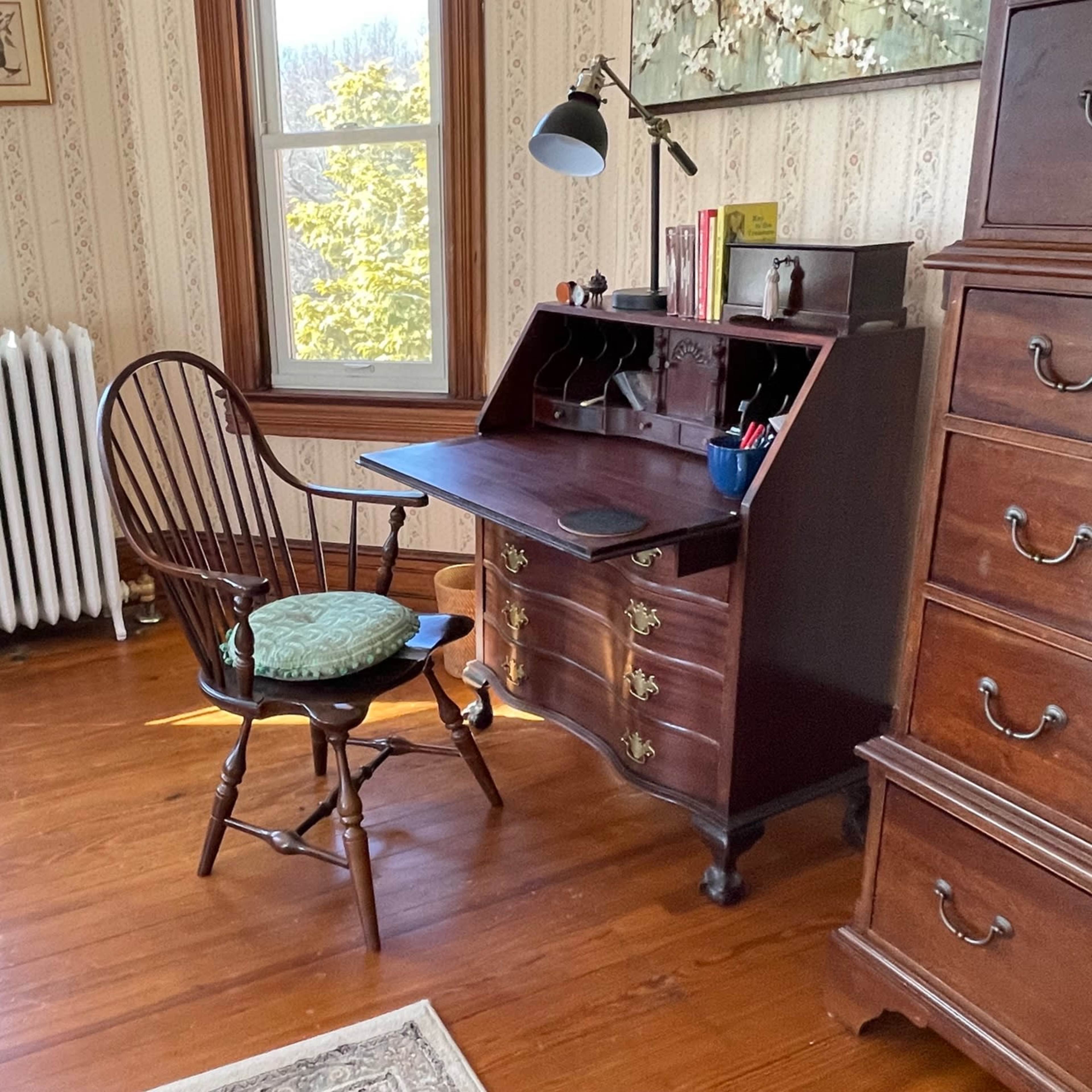 A wooden desk with a chair, a lamp, and a few items arranged on the surface, set against a wallpapered wall in a room with large windows.