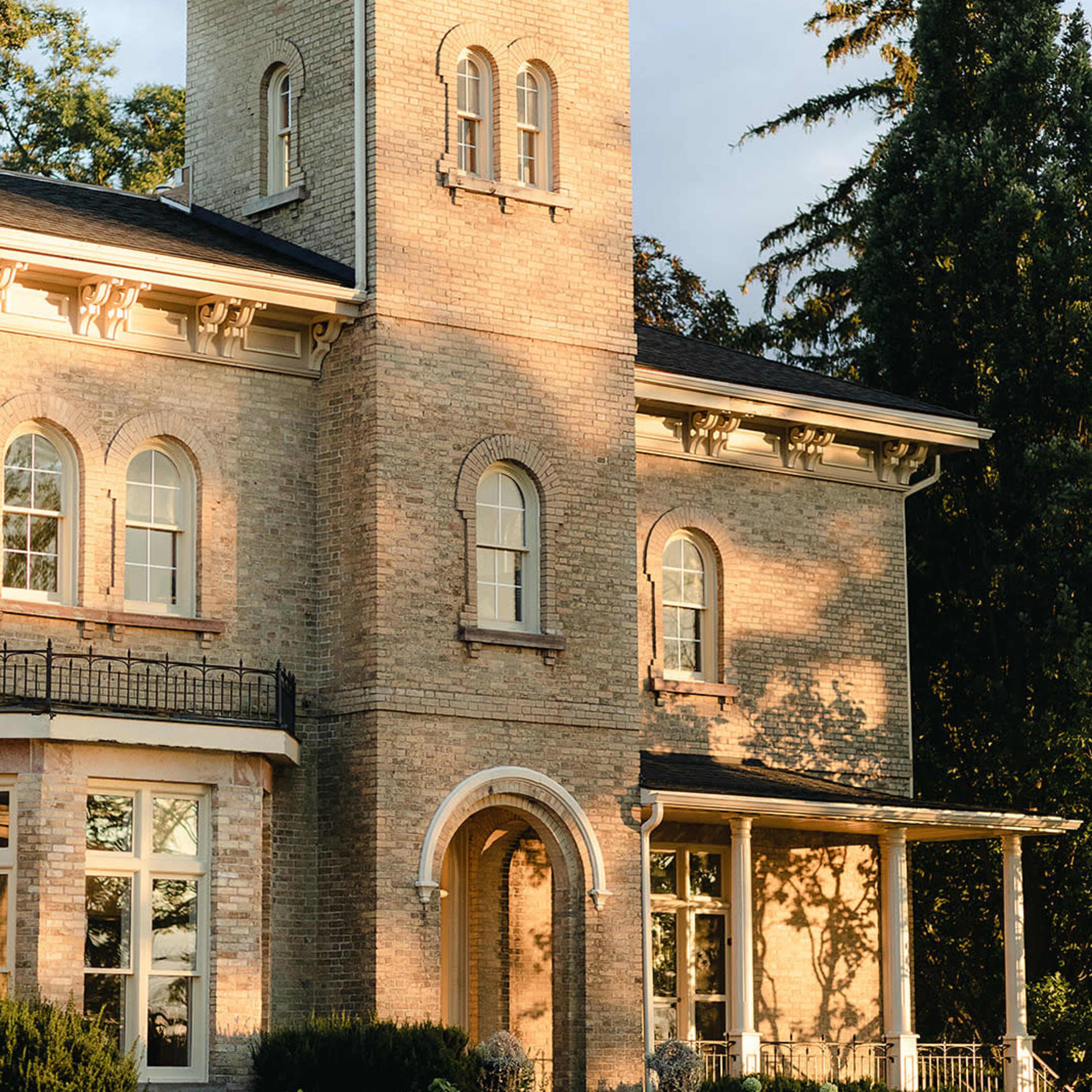 A two-story brick house with large windows, a cylindrical tower, and a front porch surrounded by greenery.