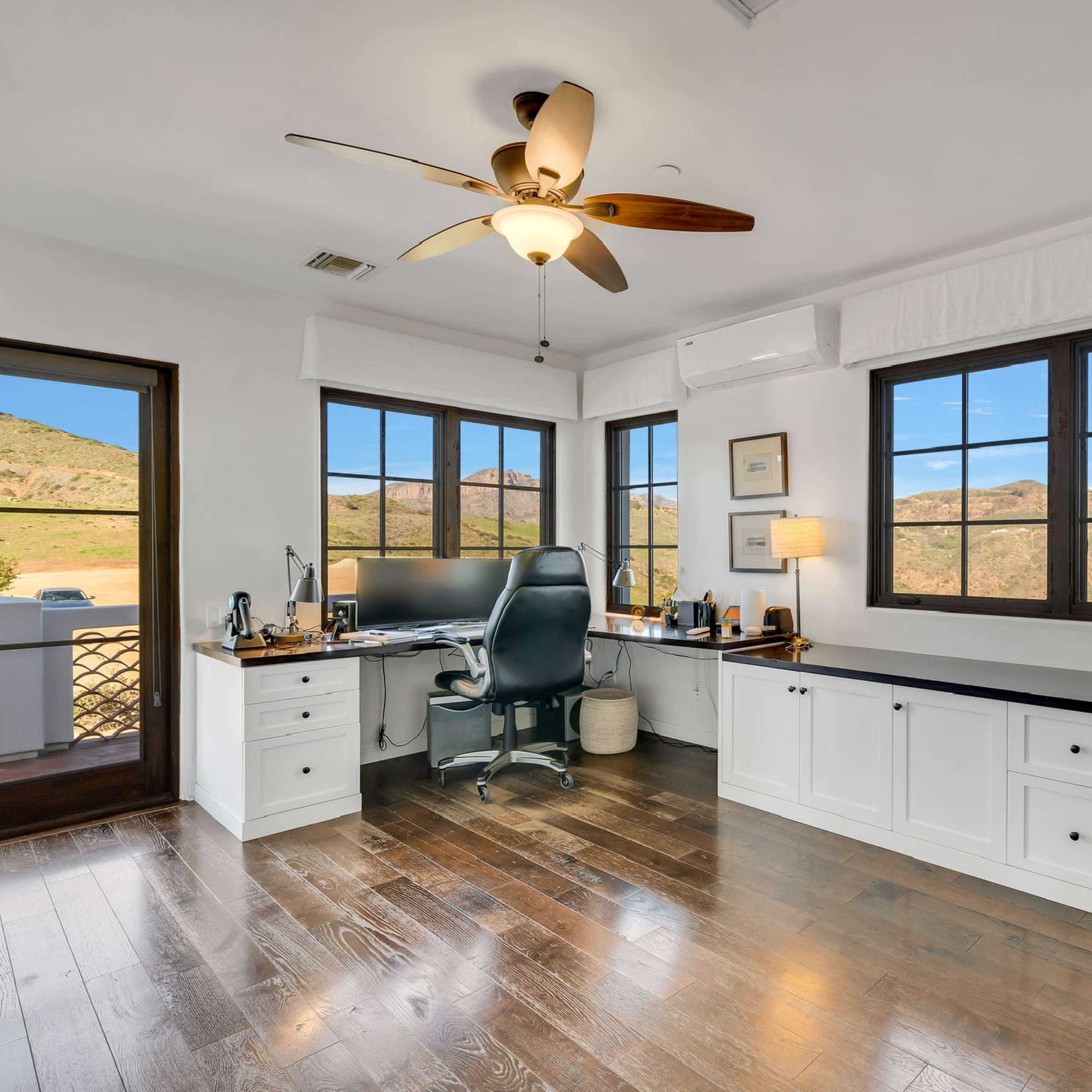 A home office features a desk with a computer, a swivel chair, and large windows overlooking a mountainous landscape.