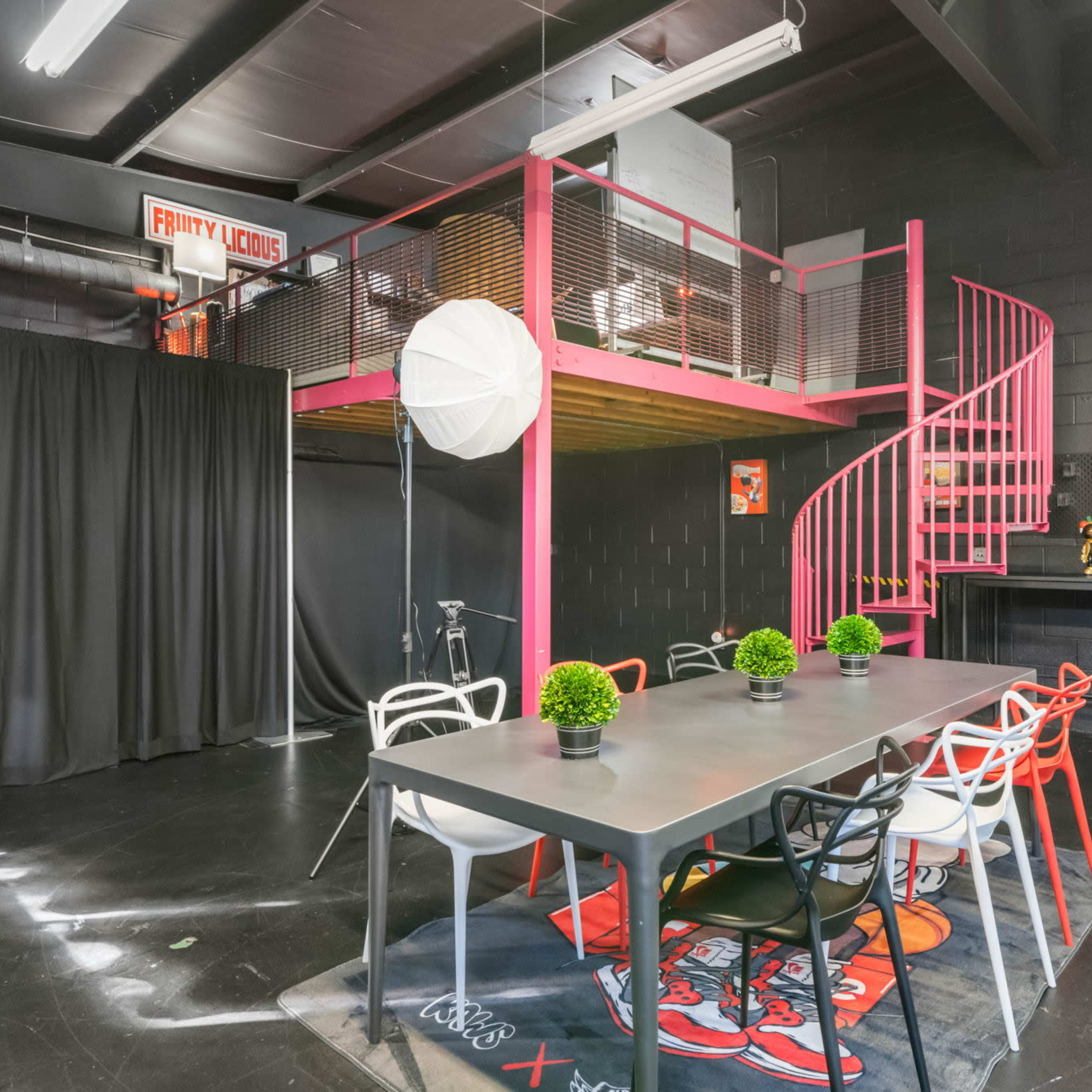 A modern studio space featuring a spiral staircase, a large table surrounded by colorful chairs, and a television on the wall, with a black curtain and potted plants in view.