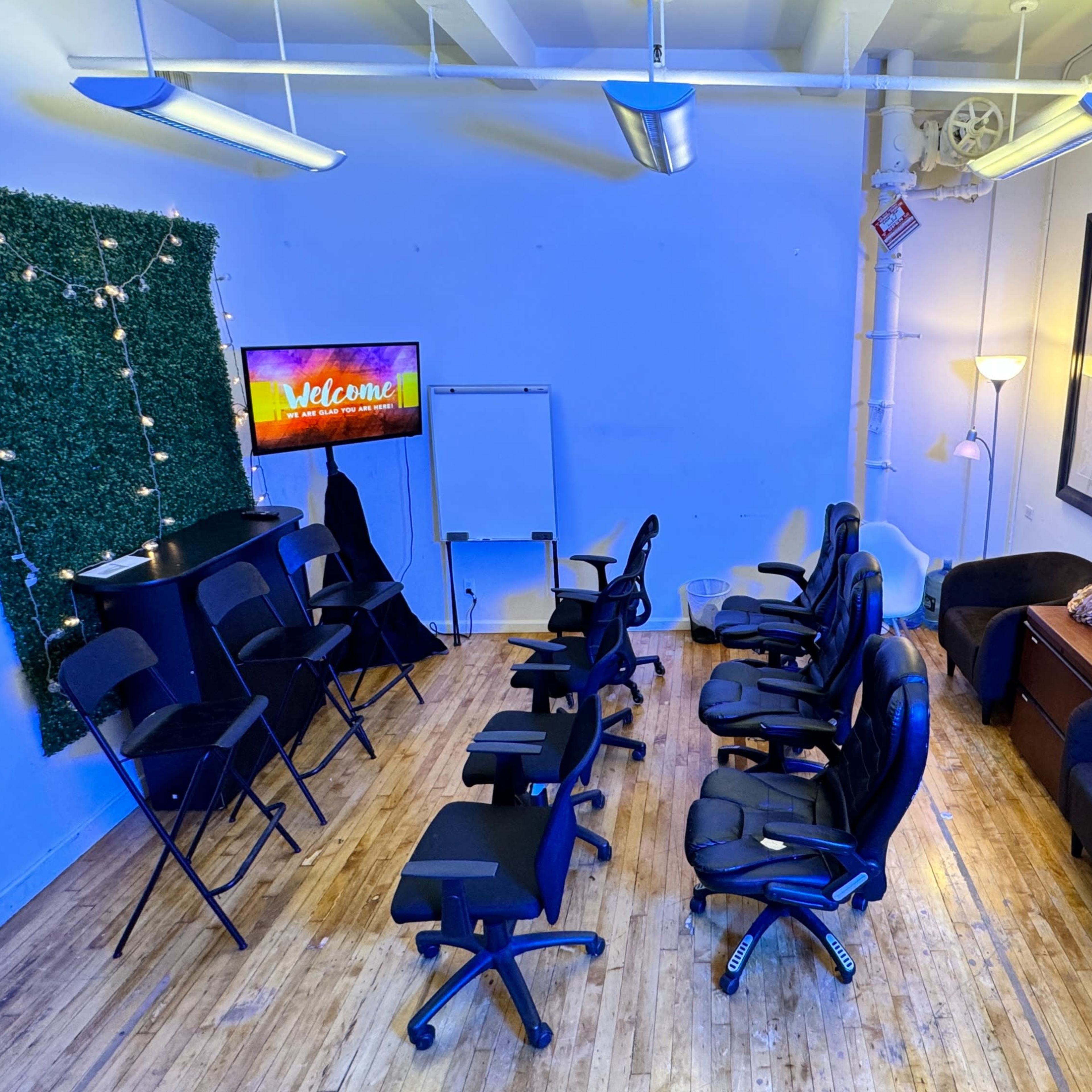 A room arranged with several black chairs, a television displaying a welcome message, and a green wall adorned with string lights.