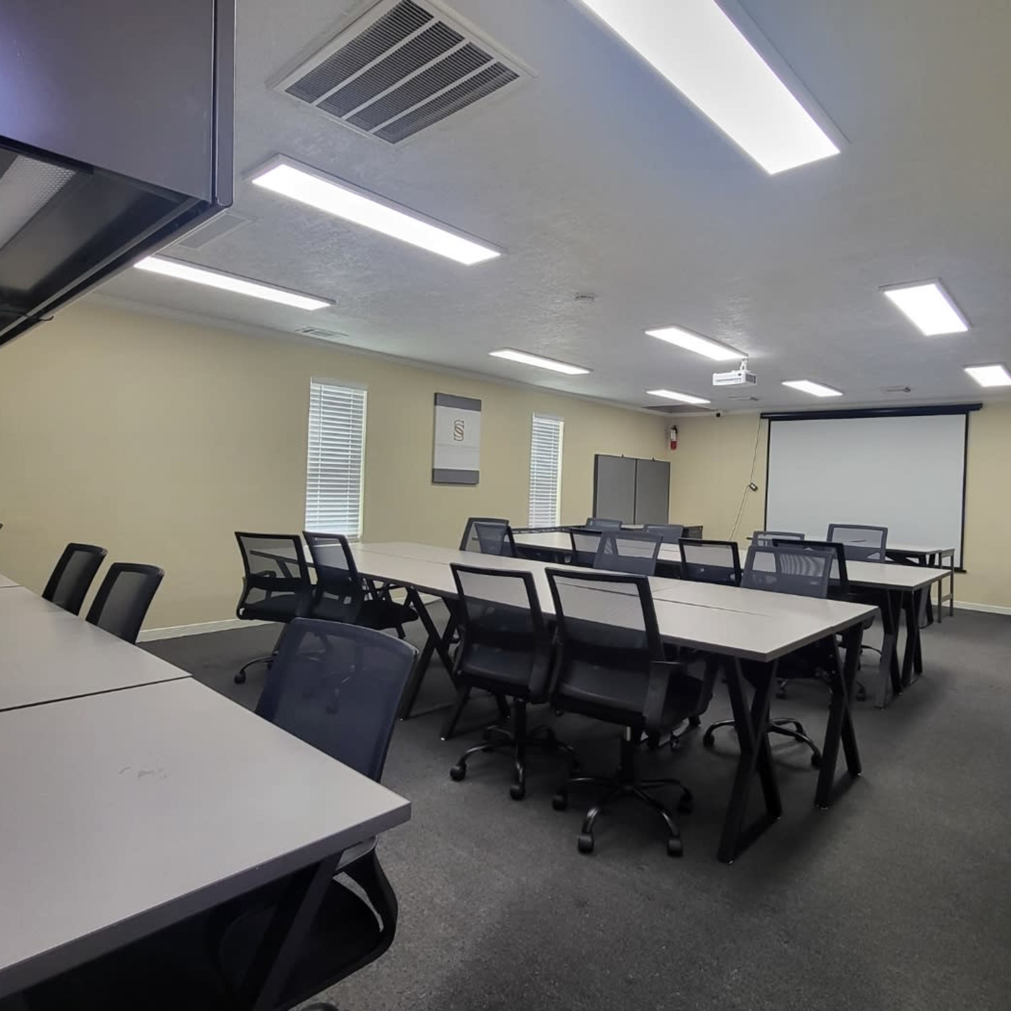 A spacious classroom features several rows of tables and chairs, a projector screen, and various educational materials on the walls.
