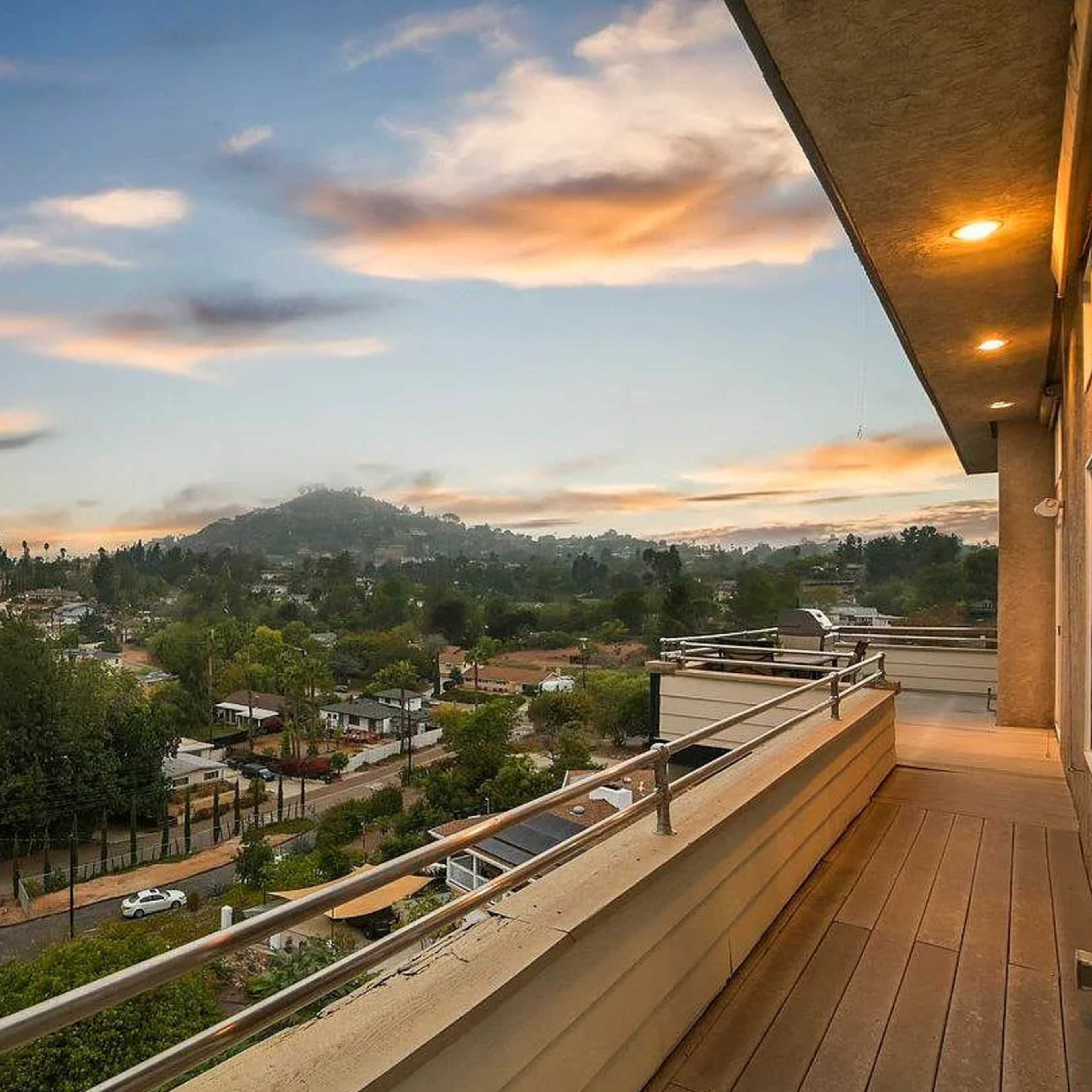 A balcony view overlooking a residential area with houses and trees, set against a backdrop of hills and a colorful sky at sunset.