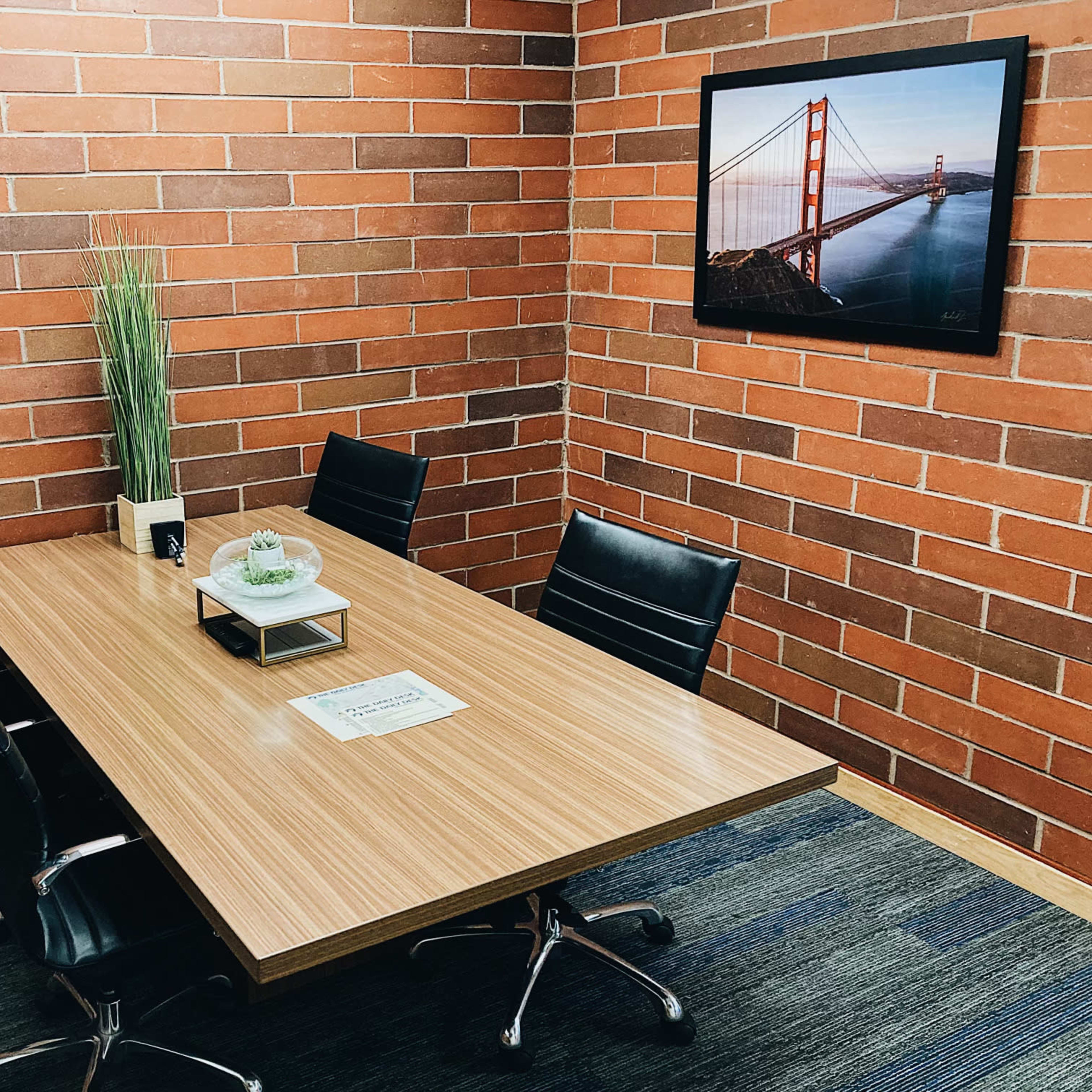 A conference room features a large wooden table surrounded by four black chairs and a framed photo of the Golden Gate Bridge on a brick wall.