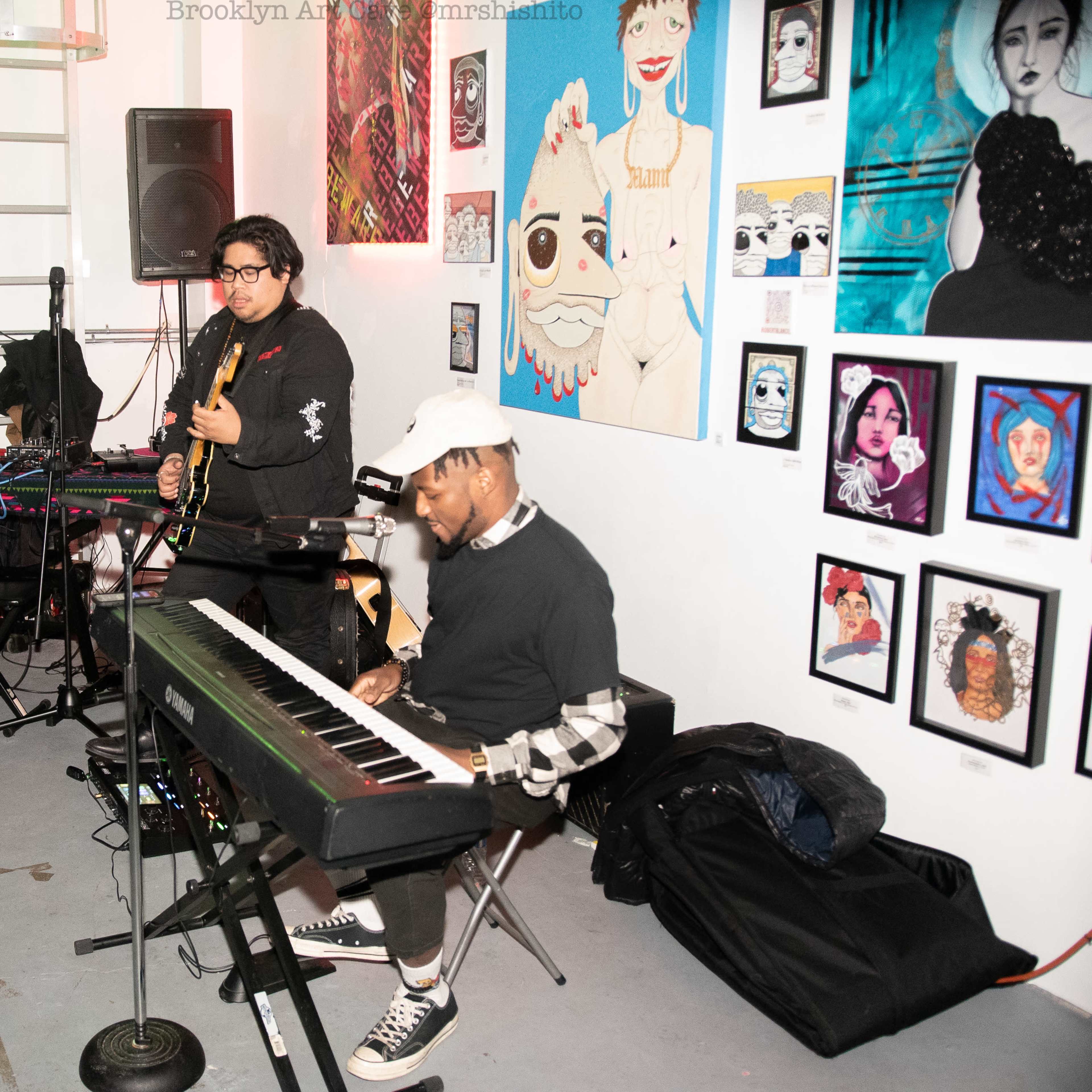 Two musicians perform in a gallery space filled with colorful artwork on the walls.