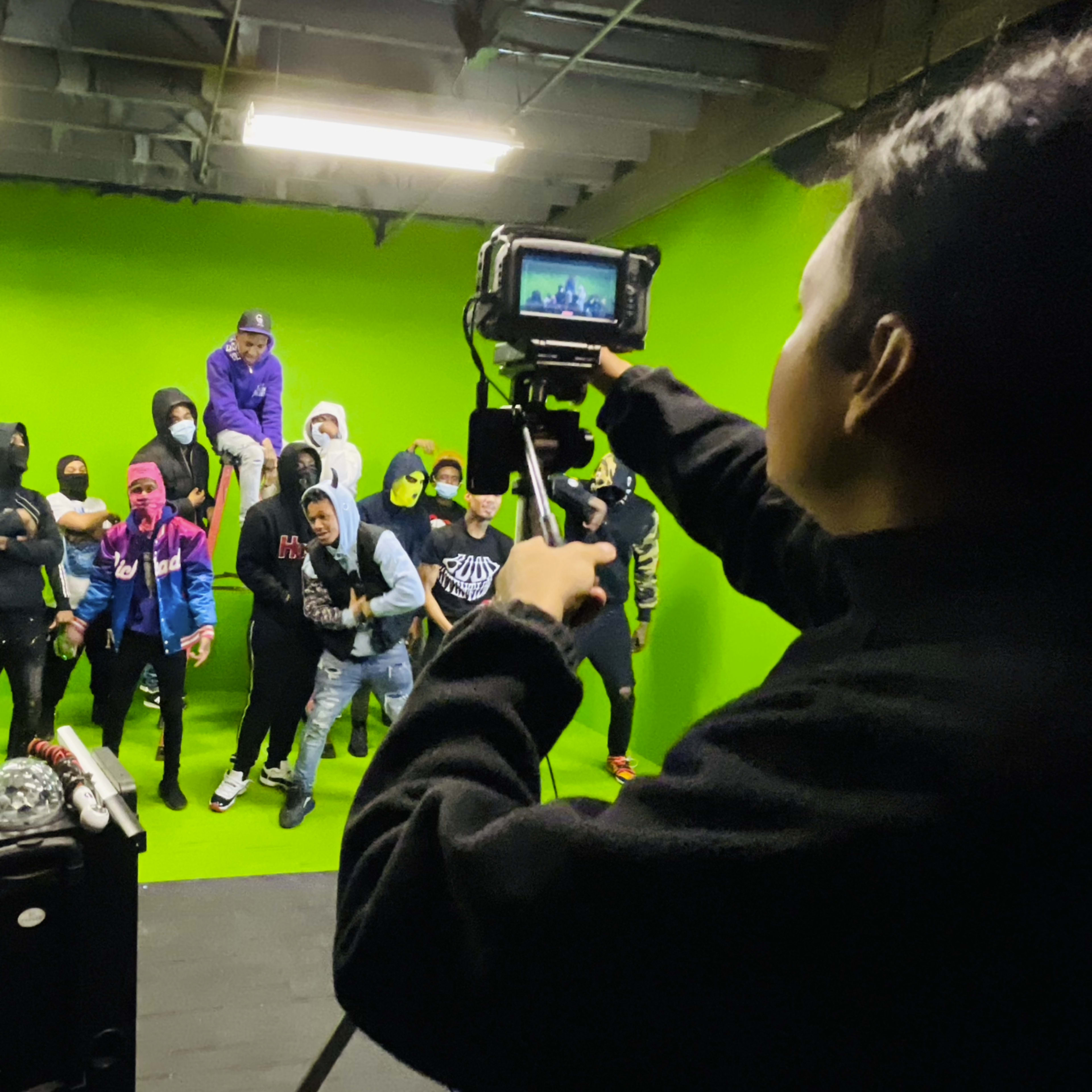 A group of individuals poses in front of a green screen while one person films them with a camera.