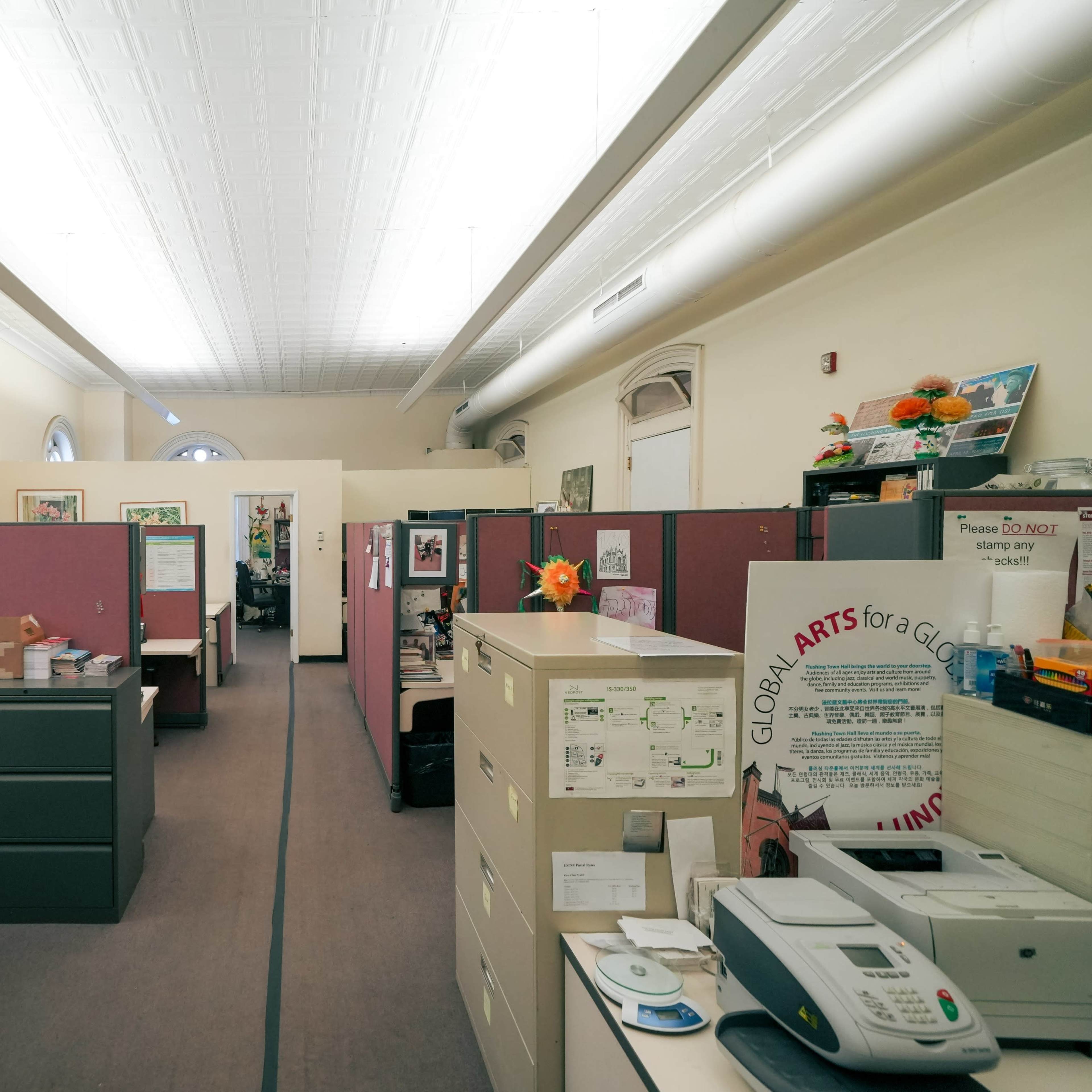 An office space with cubicles arranged along a hallway, featuring desks, filing cabinets, and various office supplies.
