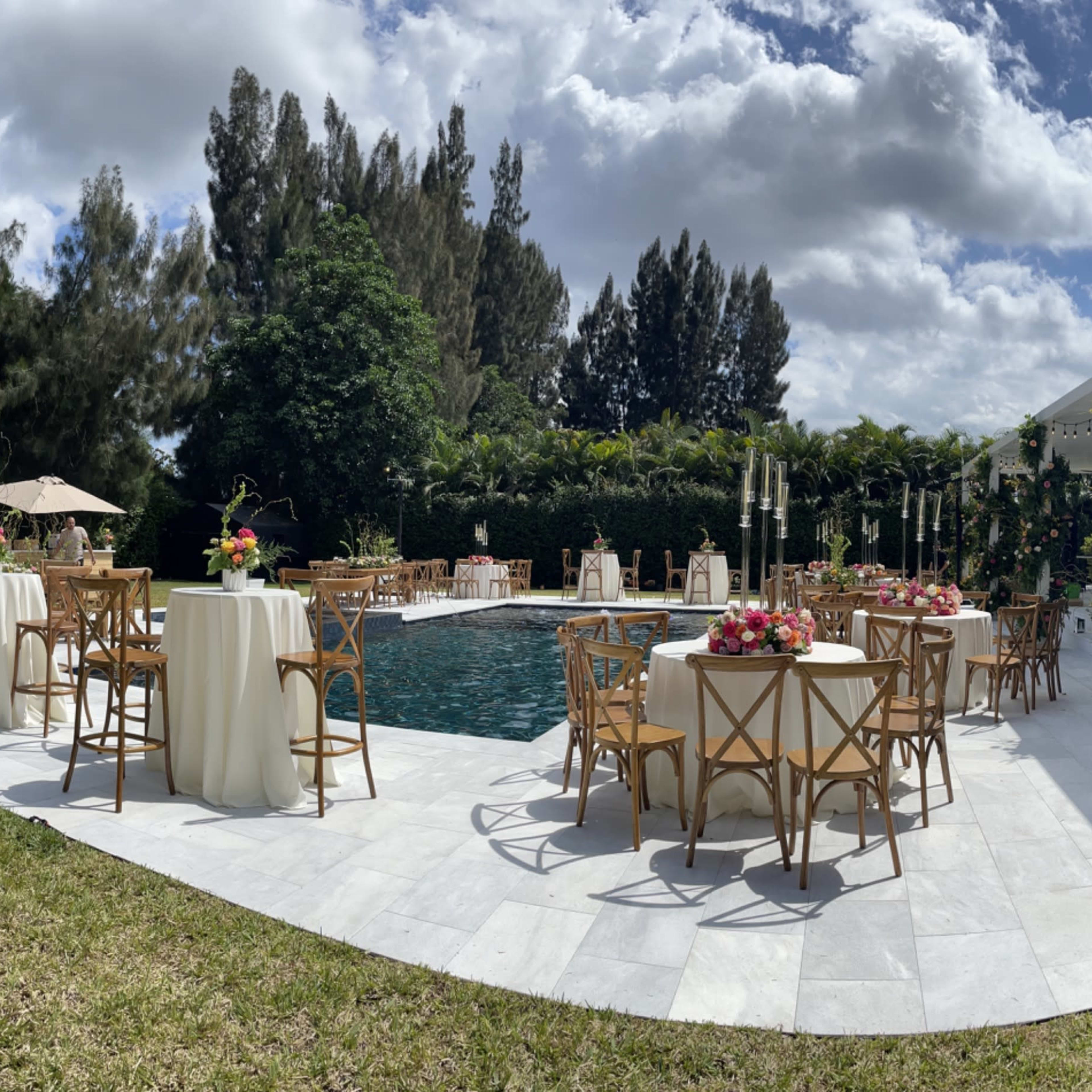 A tented outdoor event space features tables and chairs arranged around a pool, surrounded by greenery and floral decorations.