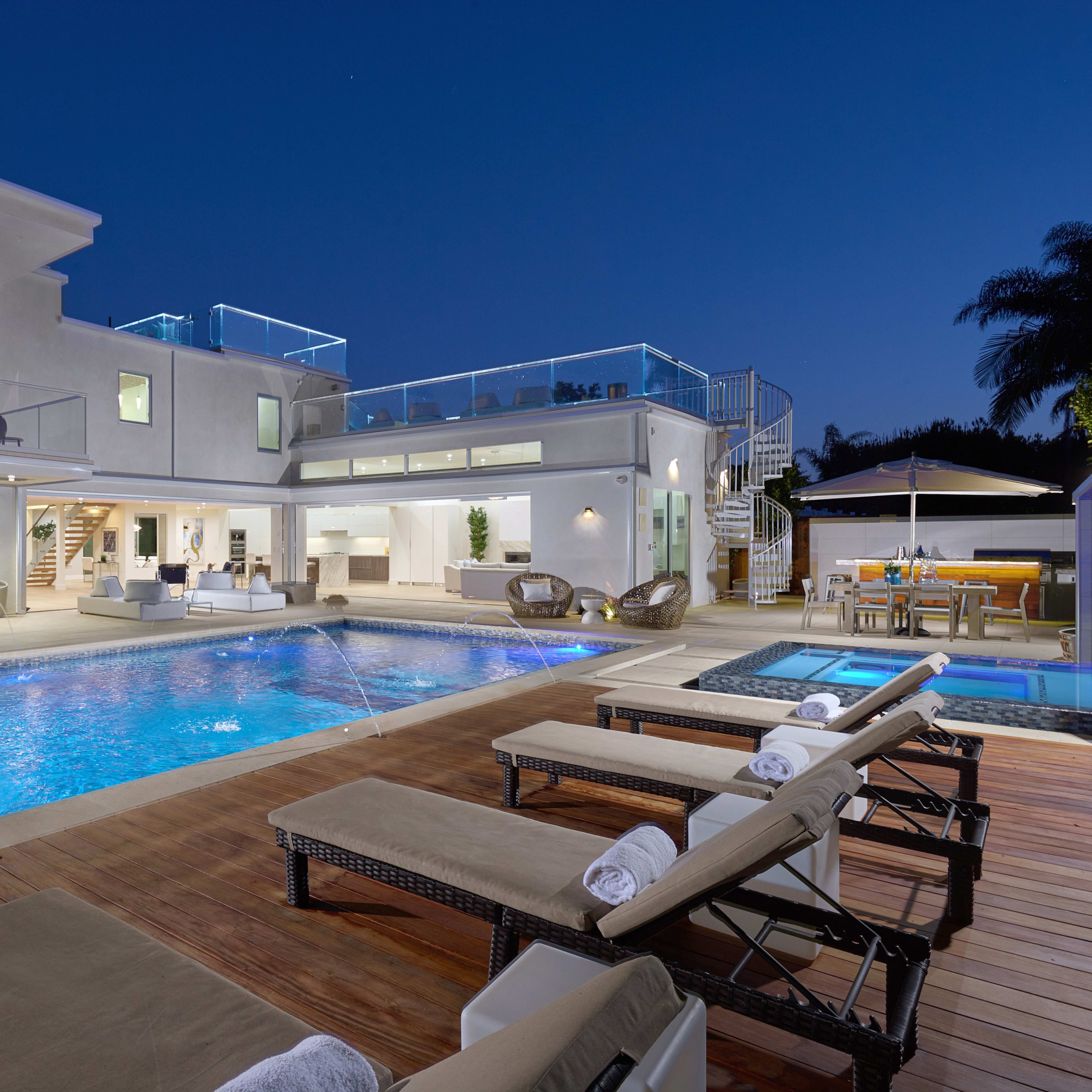 A modern two-story villa with a large swimming pool and lounge chairs, illuminated at dusk with outdoor seating areas and a hot tub.