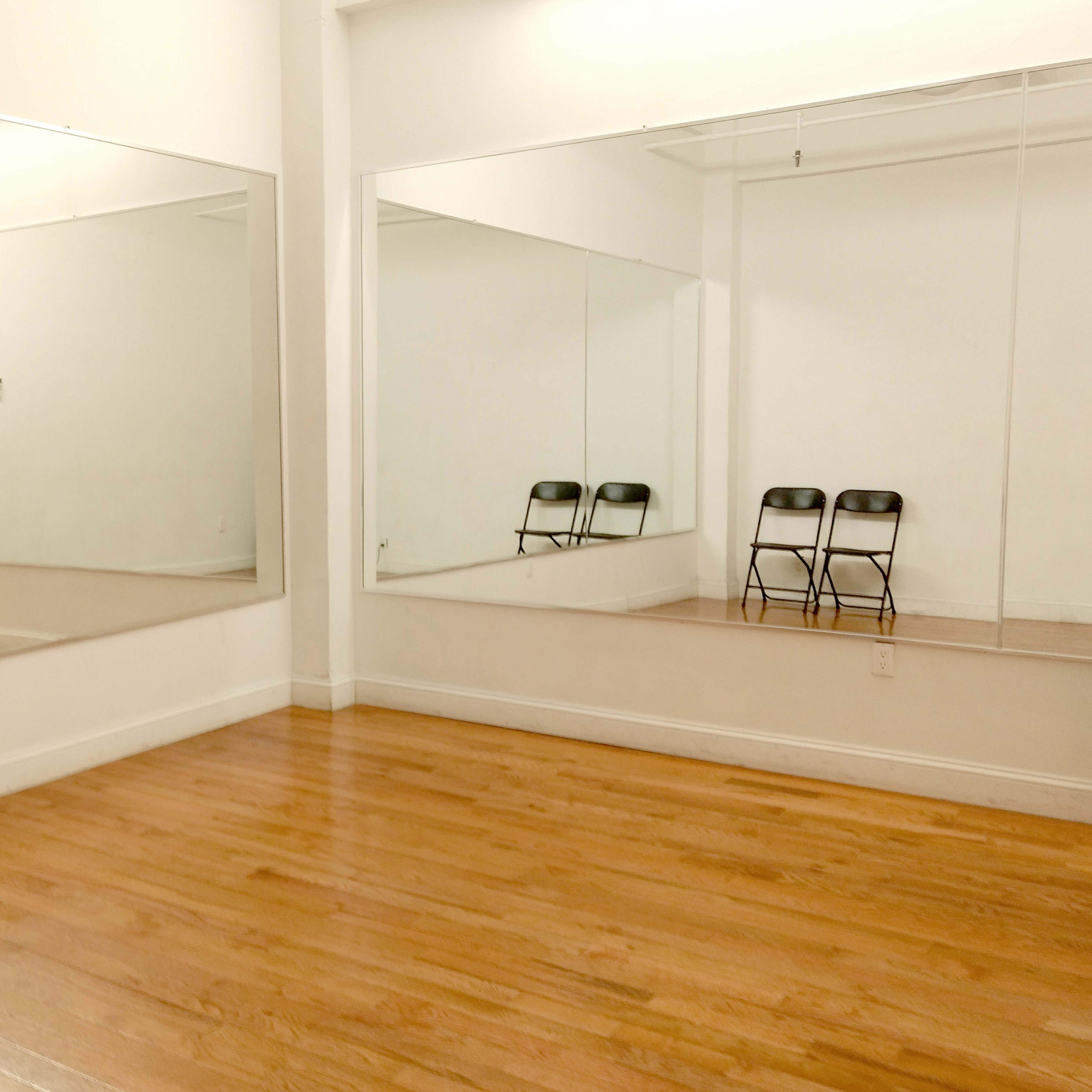 A bright room with wooden floors, featuring two large mirrors on opposite walls and three black folding chairs arranged in front of one mirror.