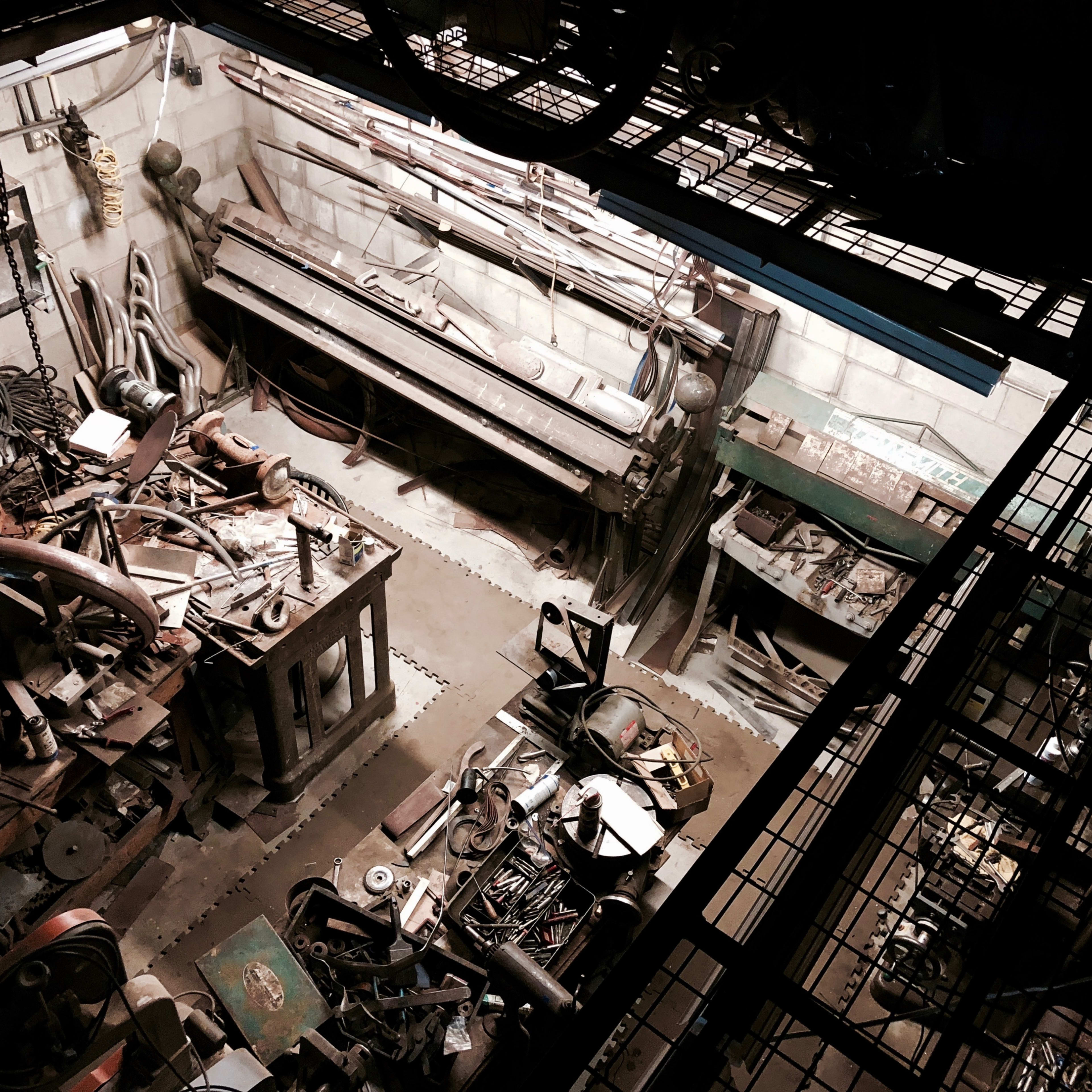 An overhead view of a cluttered industrial workshop filled with tools, machinery, and parts scattered across the floor and workbenches.
