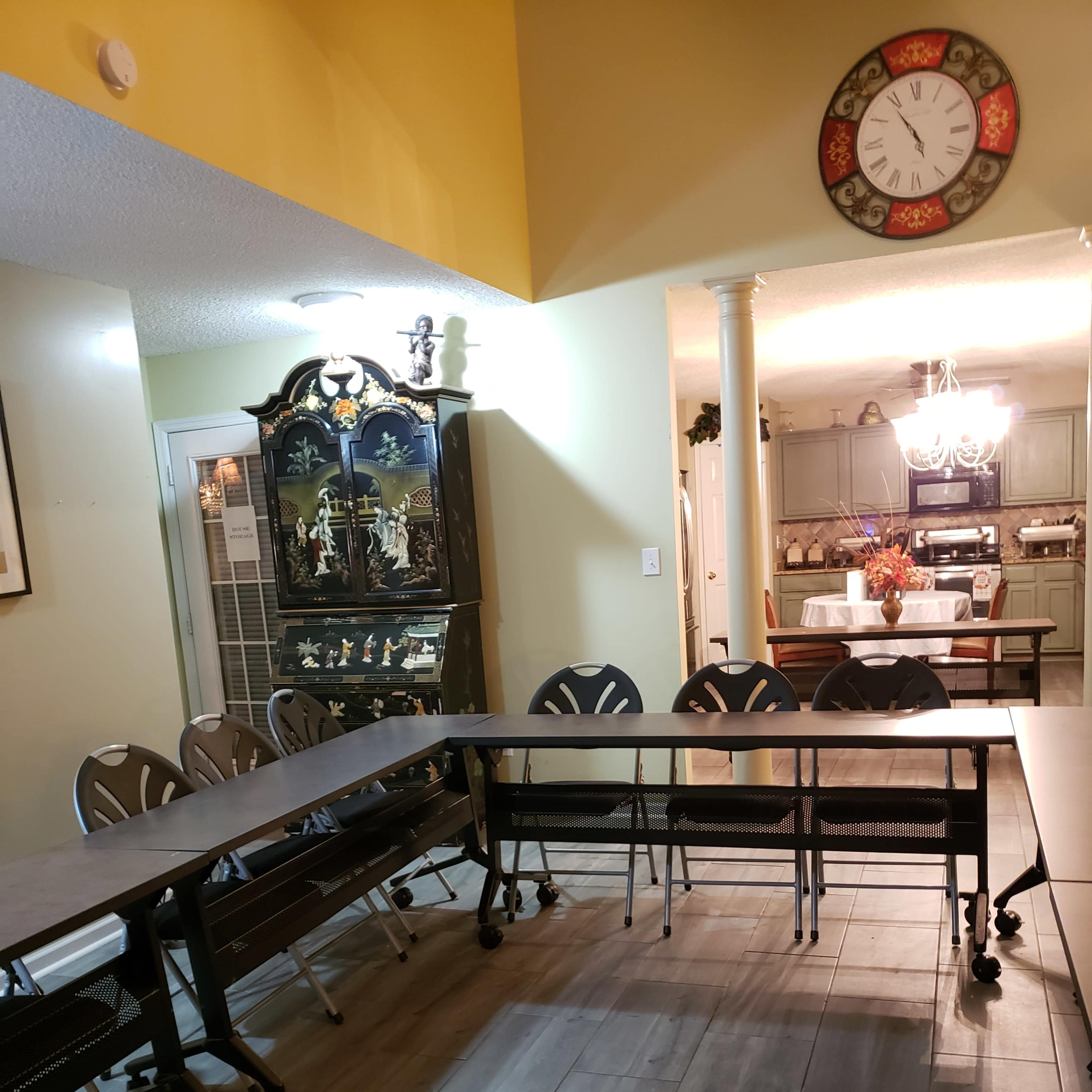 A large, rectangular table is set up with several rolling chairs around it in a well-lit room featuring a decorative cabinet and a kitchen area visible in the background.