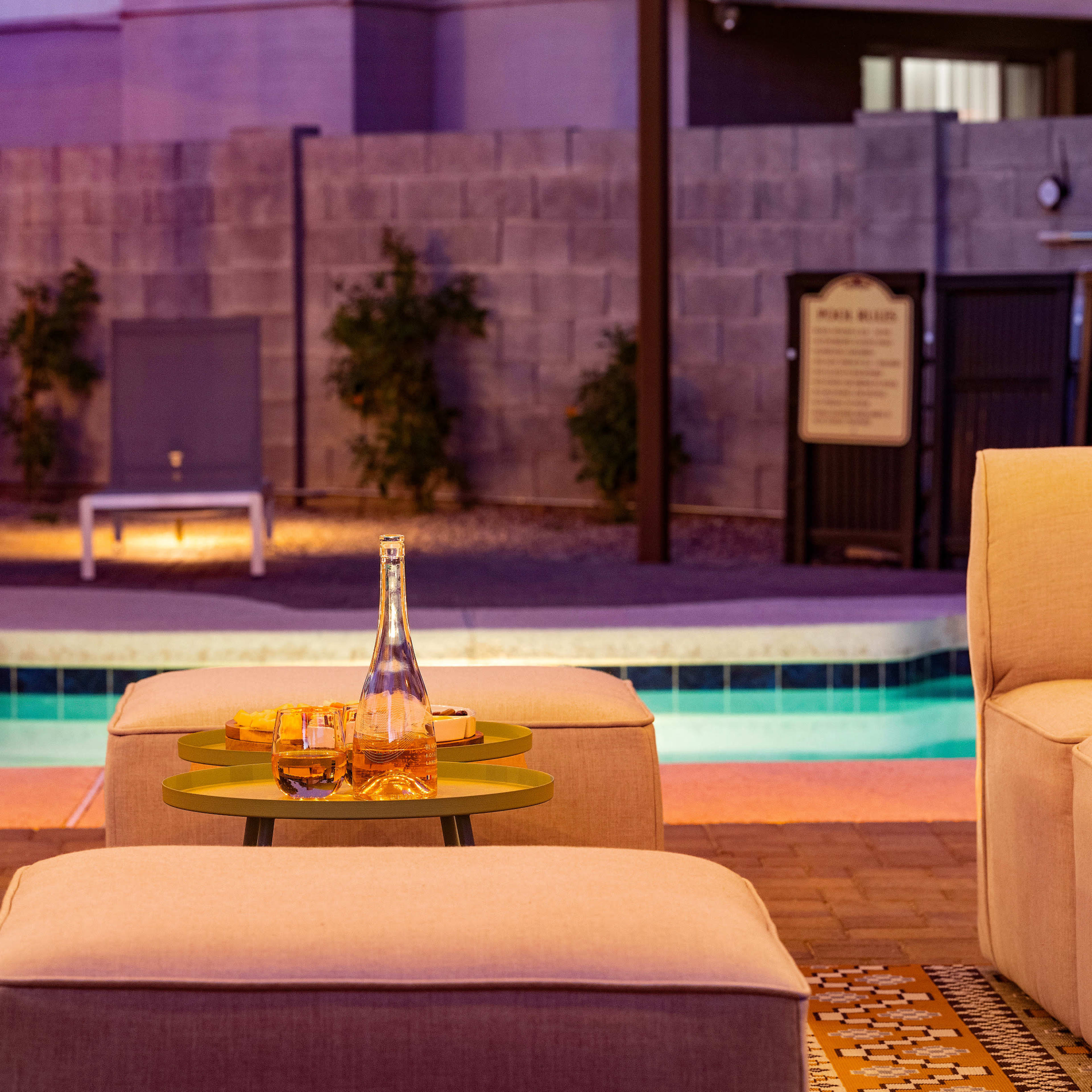 A small outdoor seating area with four light-colored chairs surrounding a green table, featuring a bottle and glasses, next to a pool illuminated by soft lighting.